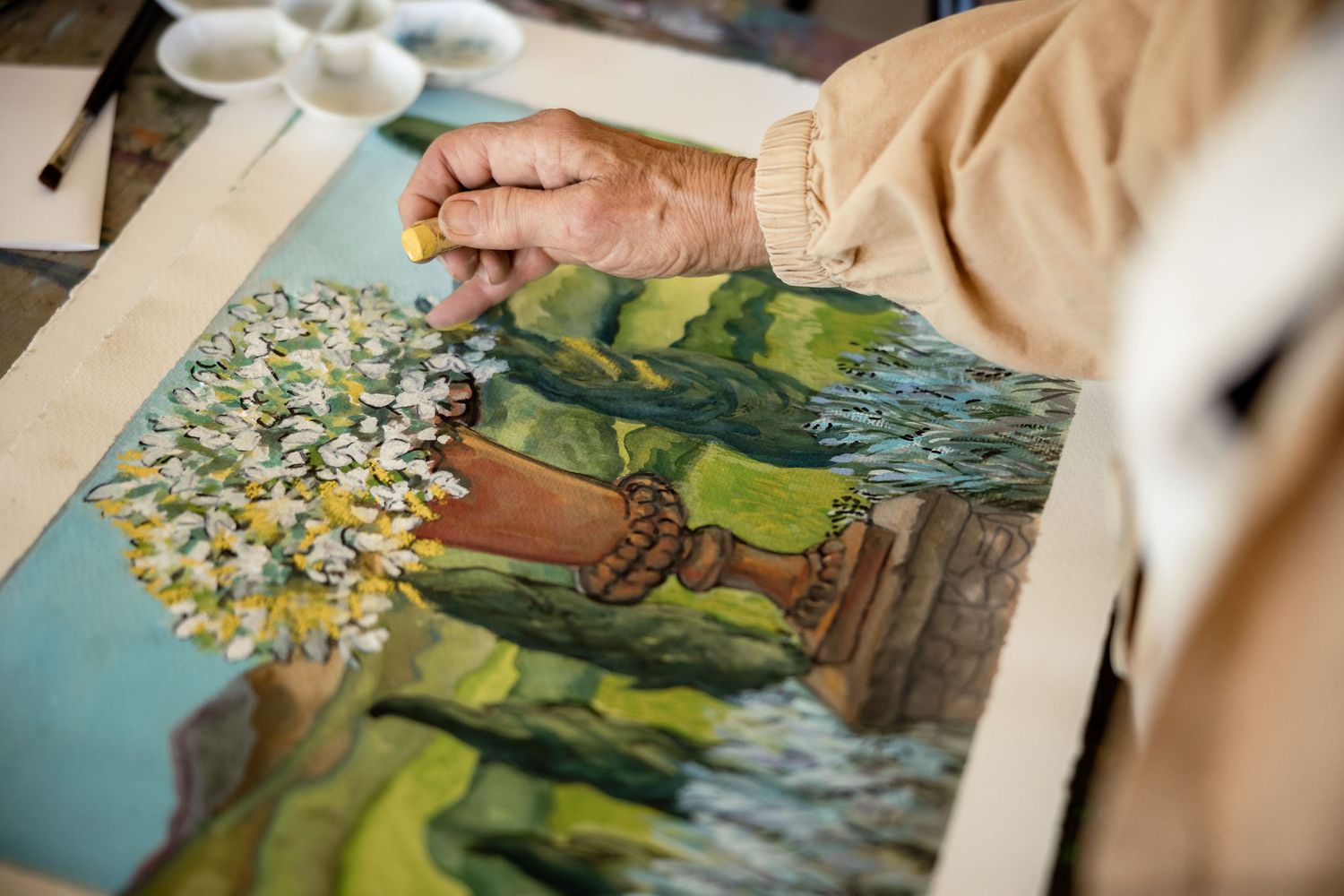 An artist's hand works on a detailed watercolor painting featuring white flowers and a green landscape with a wooden structure.
