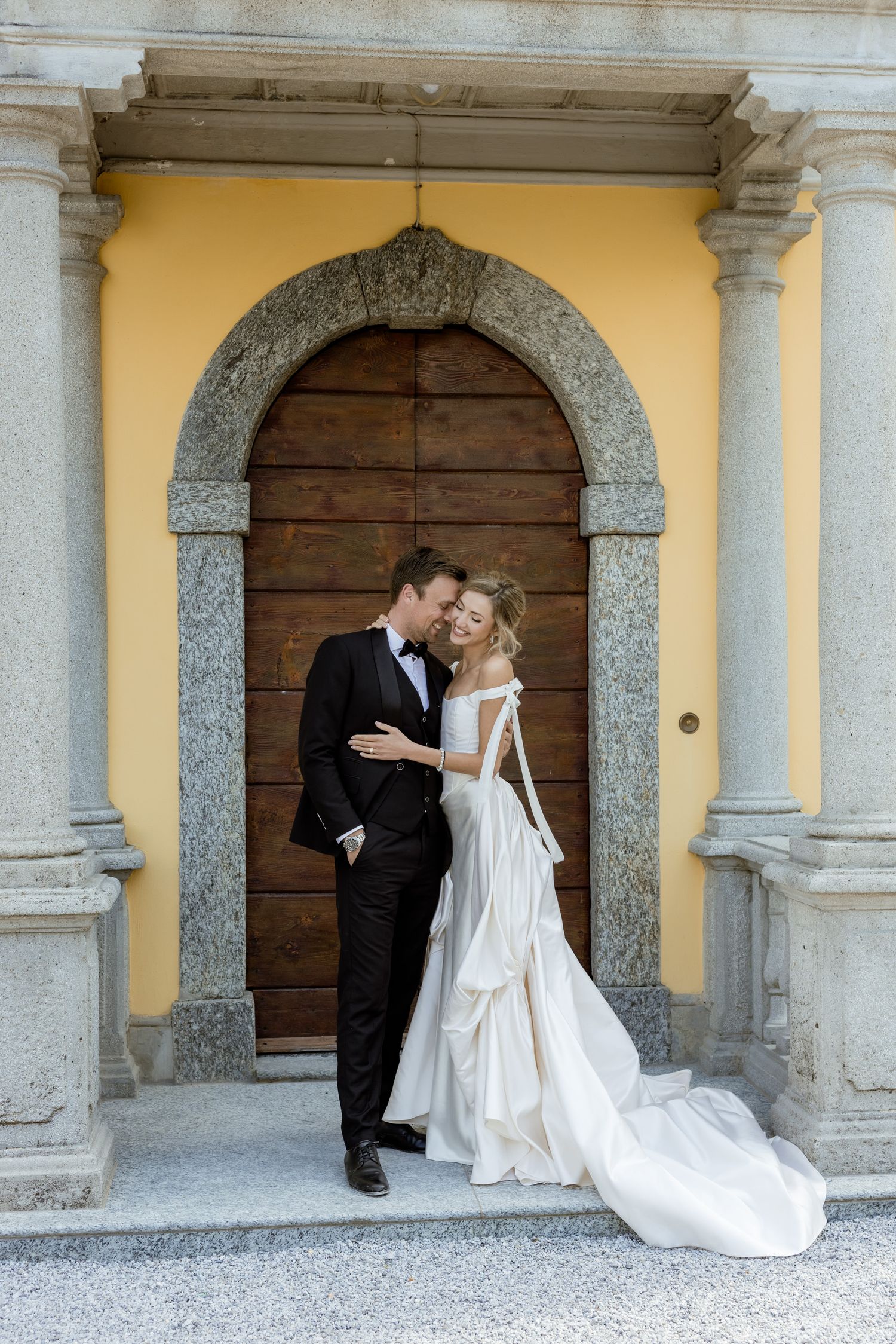 Bride and groom embracing in front of ornate arched wooden door with stone columns at elegant European venue.