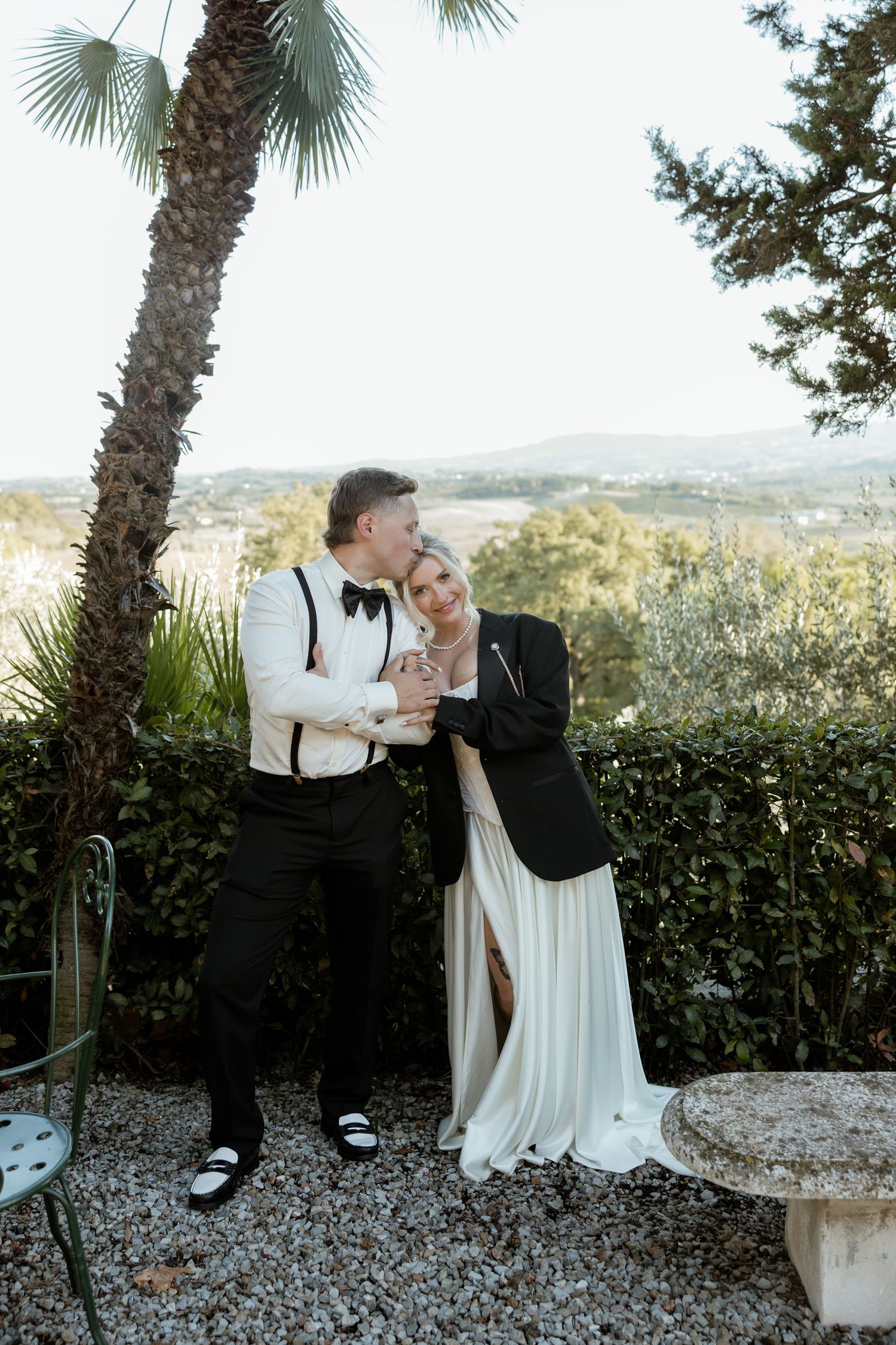 A romantic wedding couple shares tender moments under palm trees on a scenic overlook with mountain views in the background.