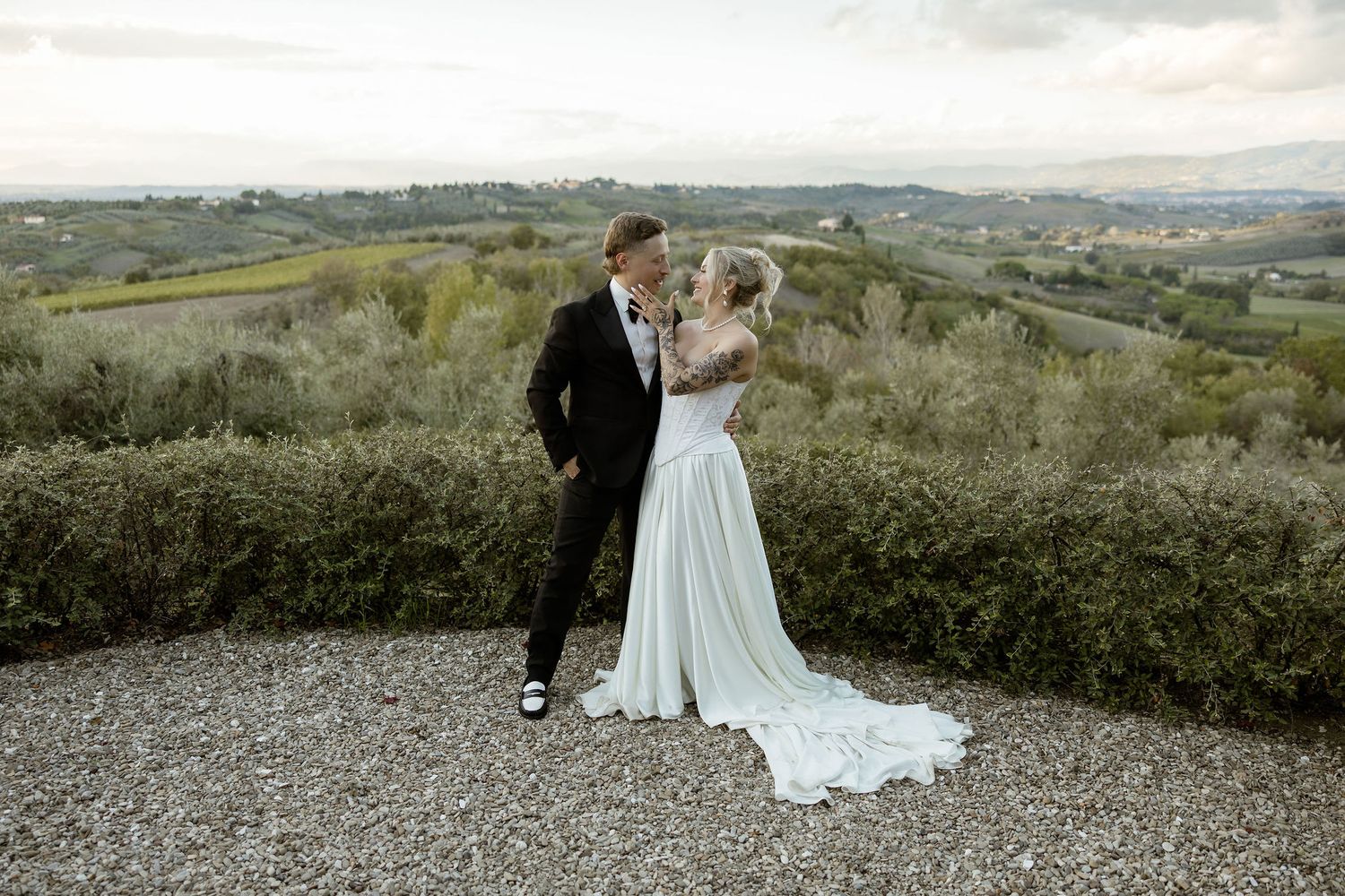 A couple stands together against a scenic countryside landscape overlooking rolling hills and distant mountains.