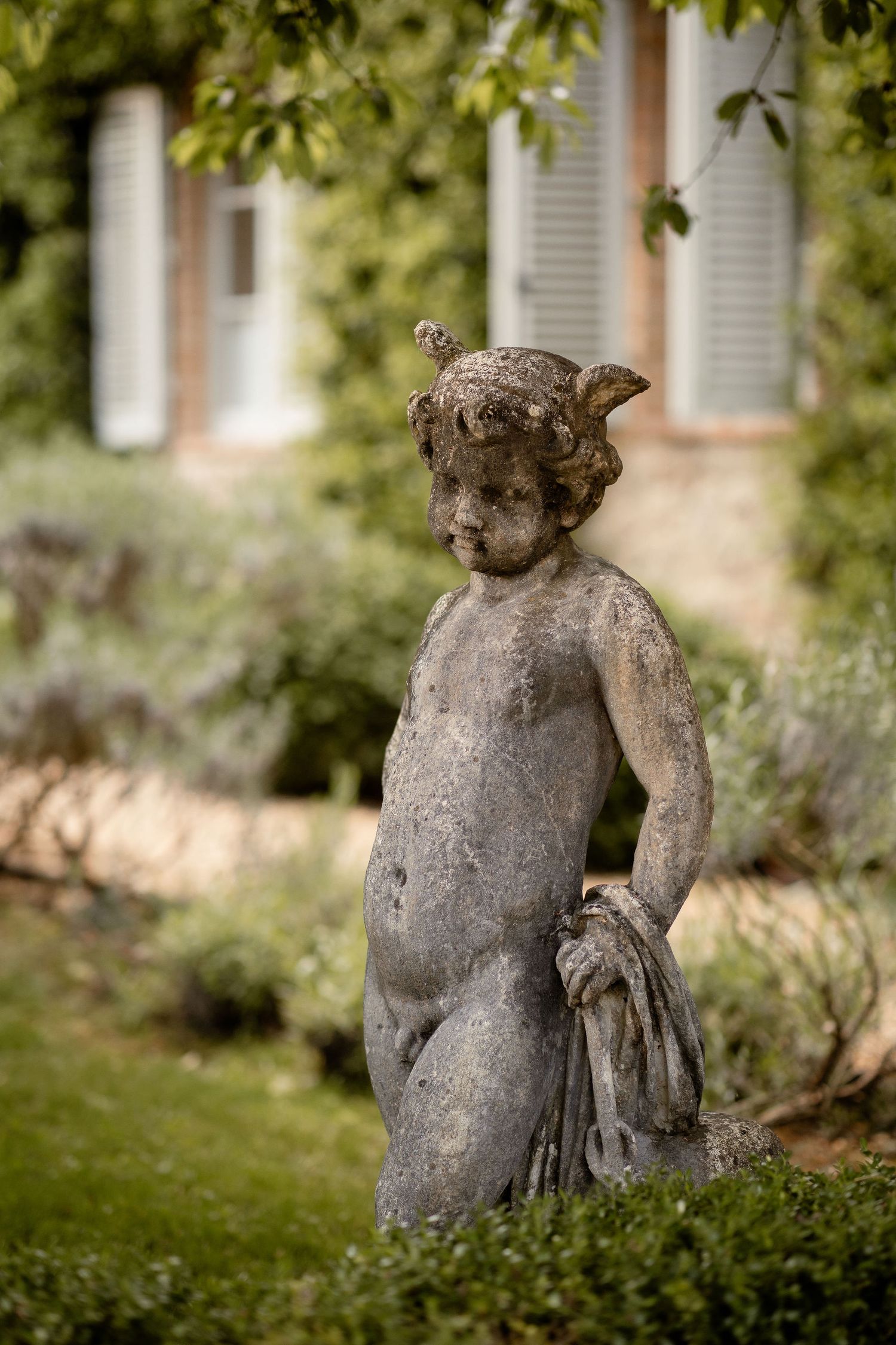 Stone cherub garden statue with decorative details standing among green bushes in a natural outdoor setting.