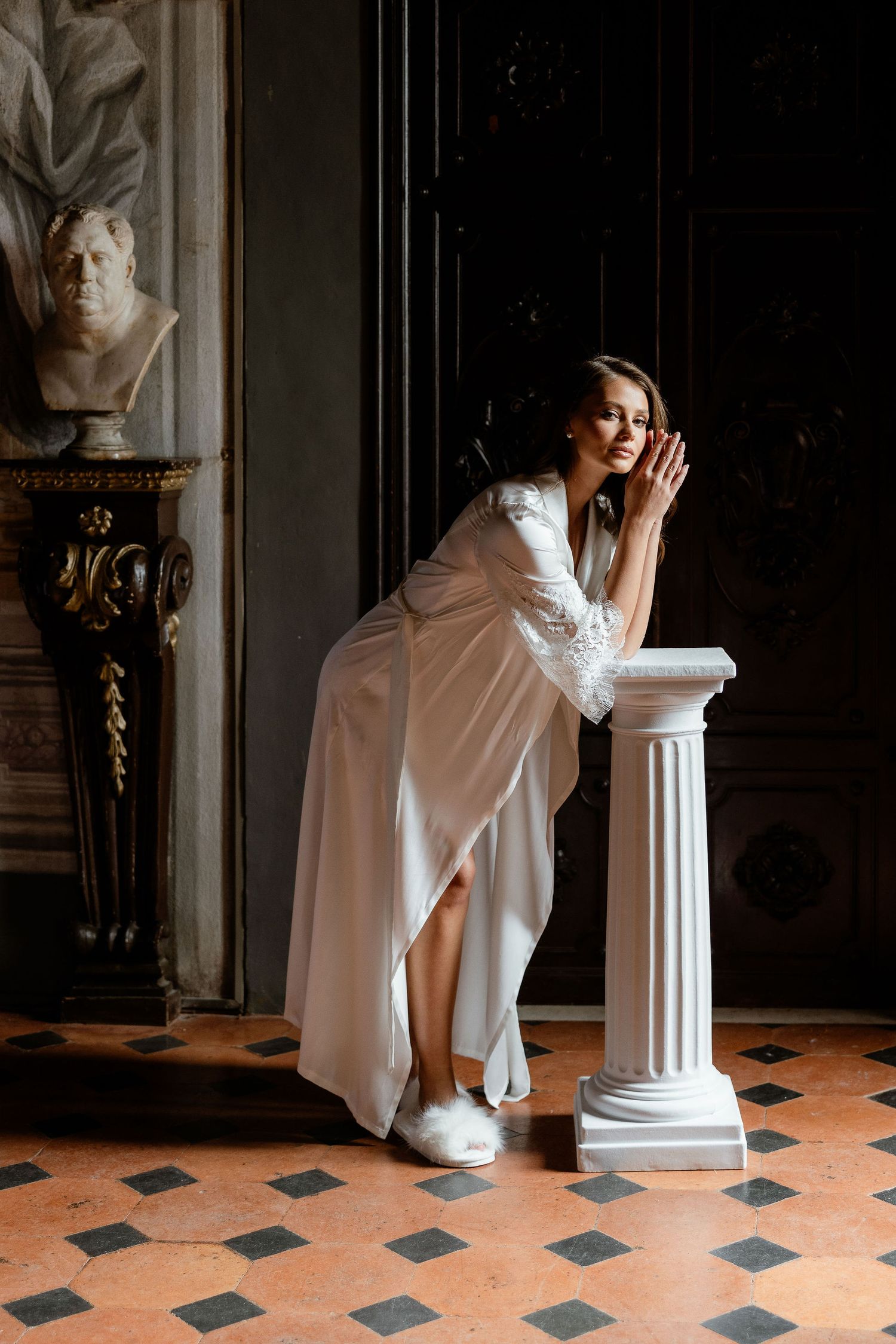 A person in a flowing white robe leans elegantly against a classical column in a room with terracotta tile floors.