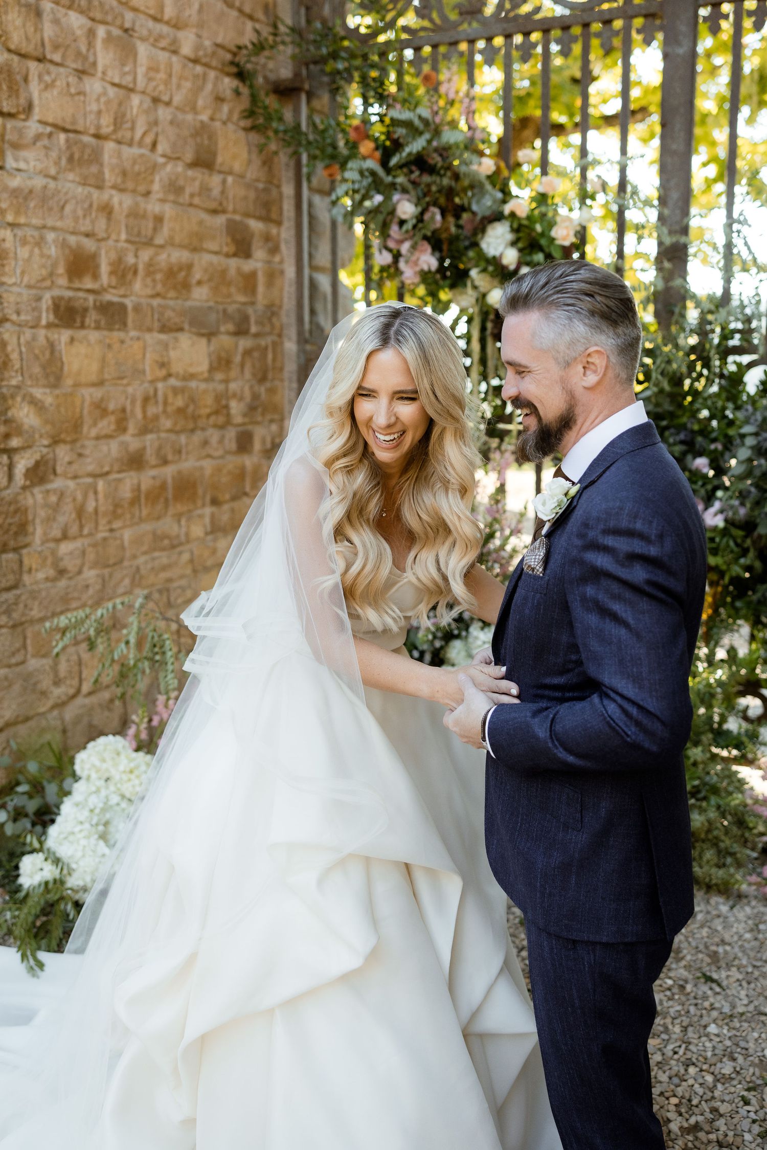 A bride and groom share joyful candid moments together outdoors against a stone wall on their wedding day.