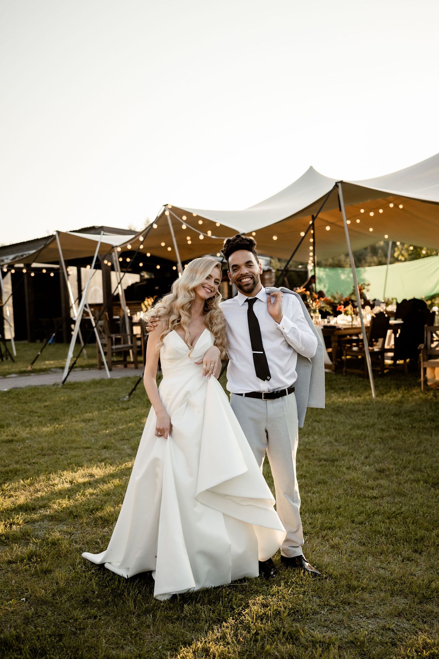 Newlyweds share intimate moment under string-lit reception tent at sunset.