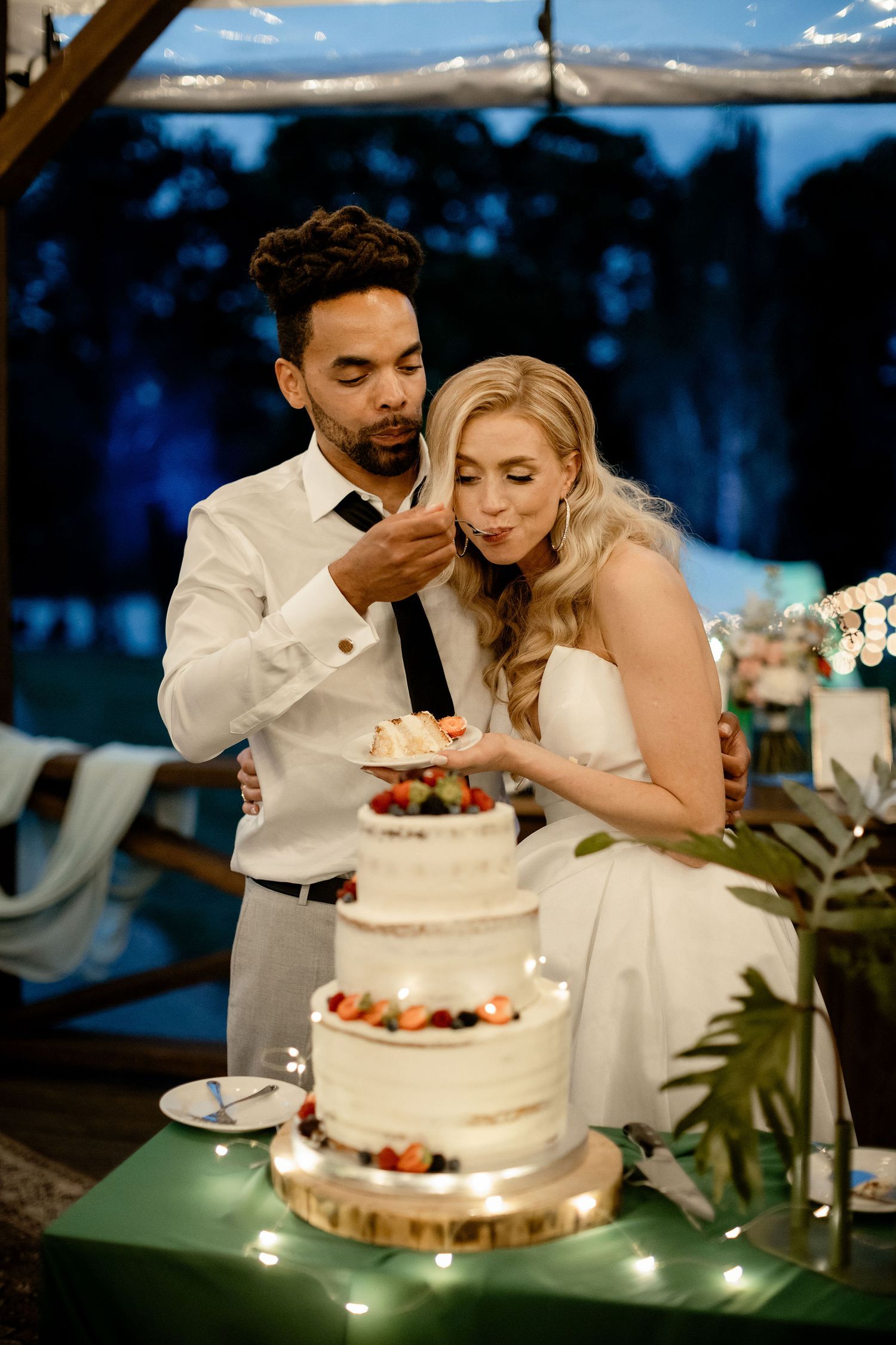 A couple feeds each other wedding cake at their outdoor reception with a three-tiered white cake decorated with berries.