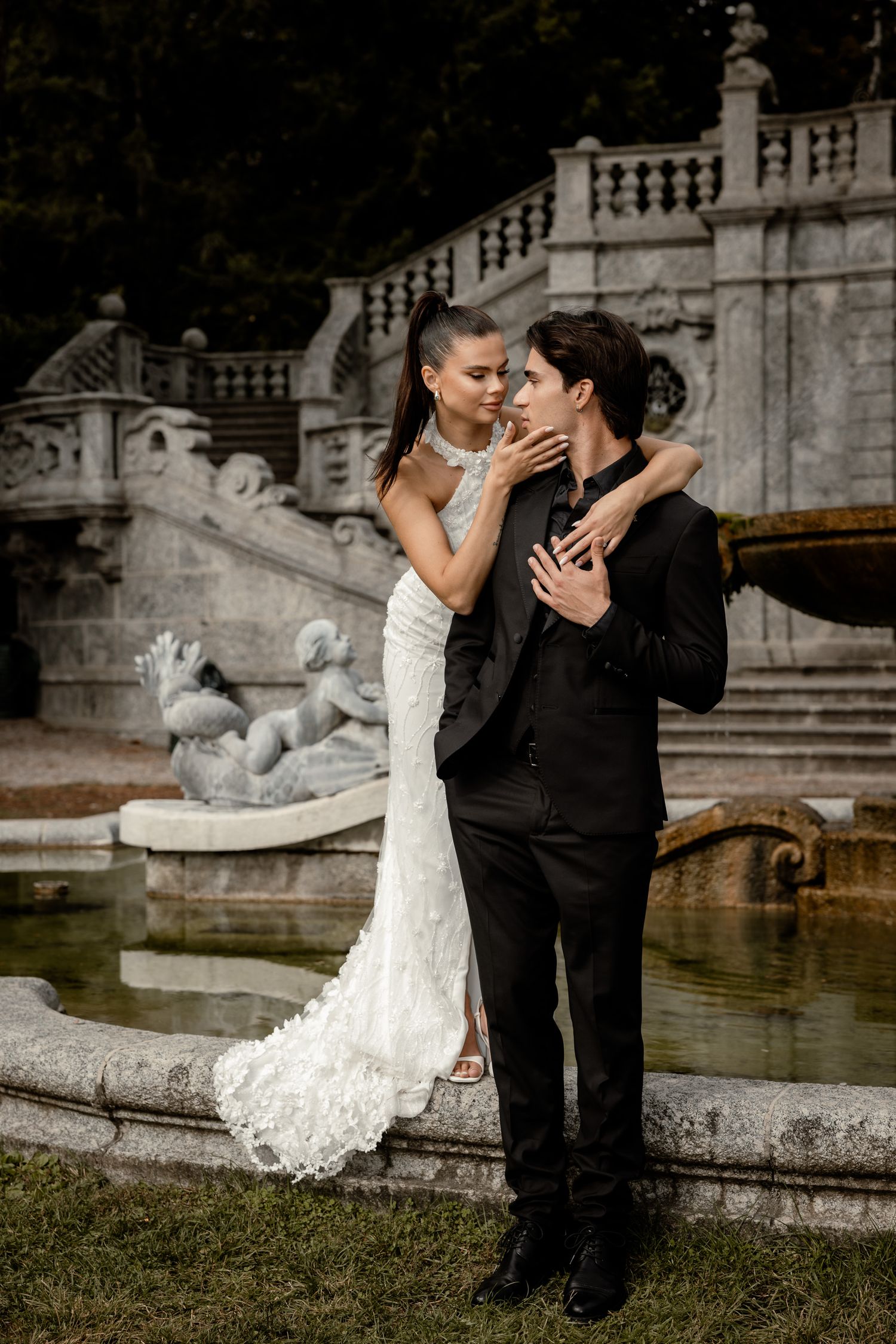 Romantic wedding couple embraces by ornate fountain in formal garden setting.