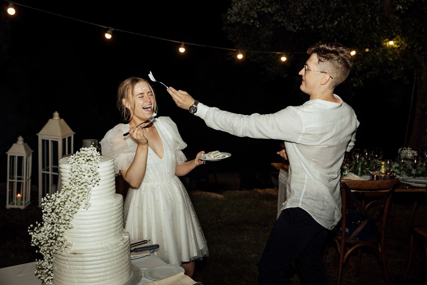 A couple shares a playful moment under string lights at an outdoor nighttime celebration.