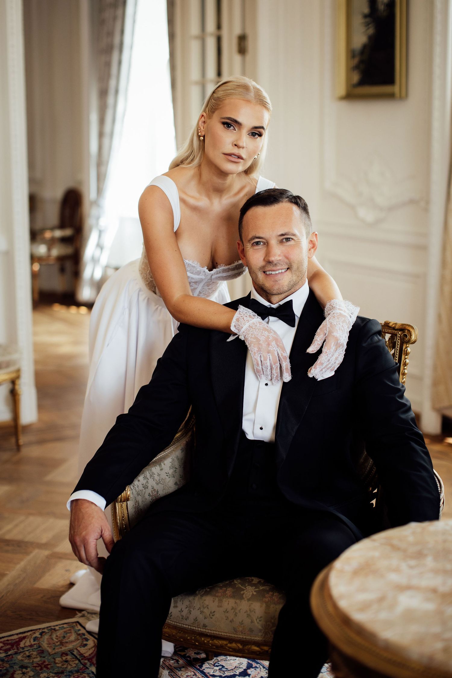 Elegant couple in formal attire pose together on vintage chair in luxurious room with white curtains and ornate decor.