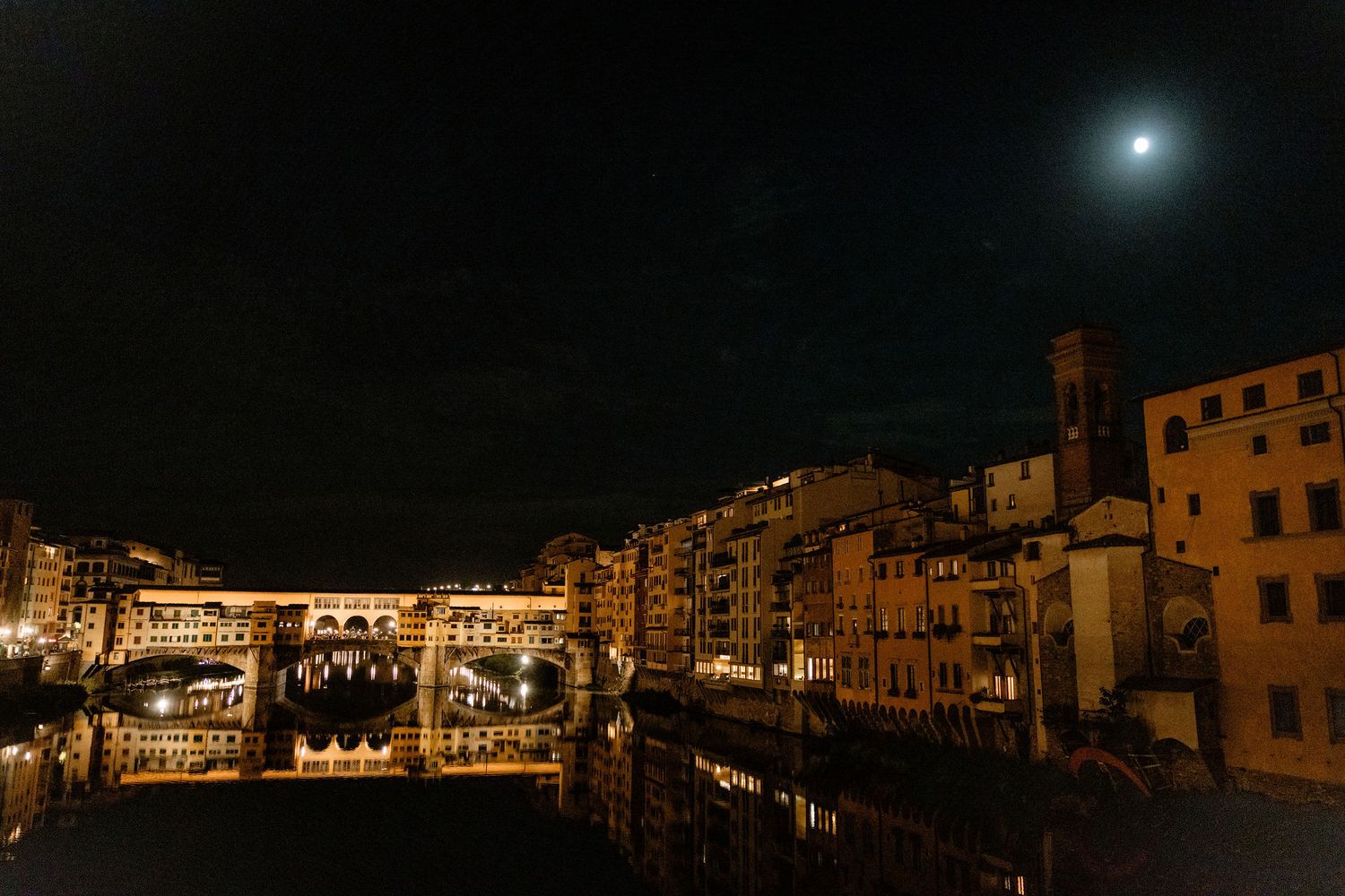 A nighttime view of Florence's illuminated Ponte Vecchio bridge with a full moon glowing above the Arno River.