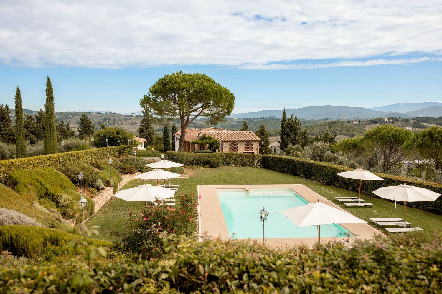 Luxurious resort swimming pool surrounded by umbrellas and loungers in Tuscan landscape.