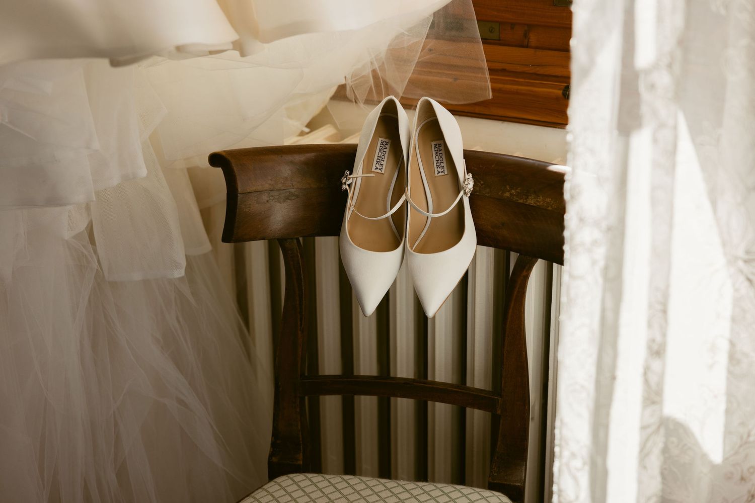 White wedding dress and heels displayed near a window with soft natural light streaming through sheer curtains.
