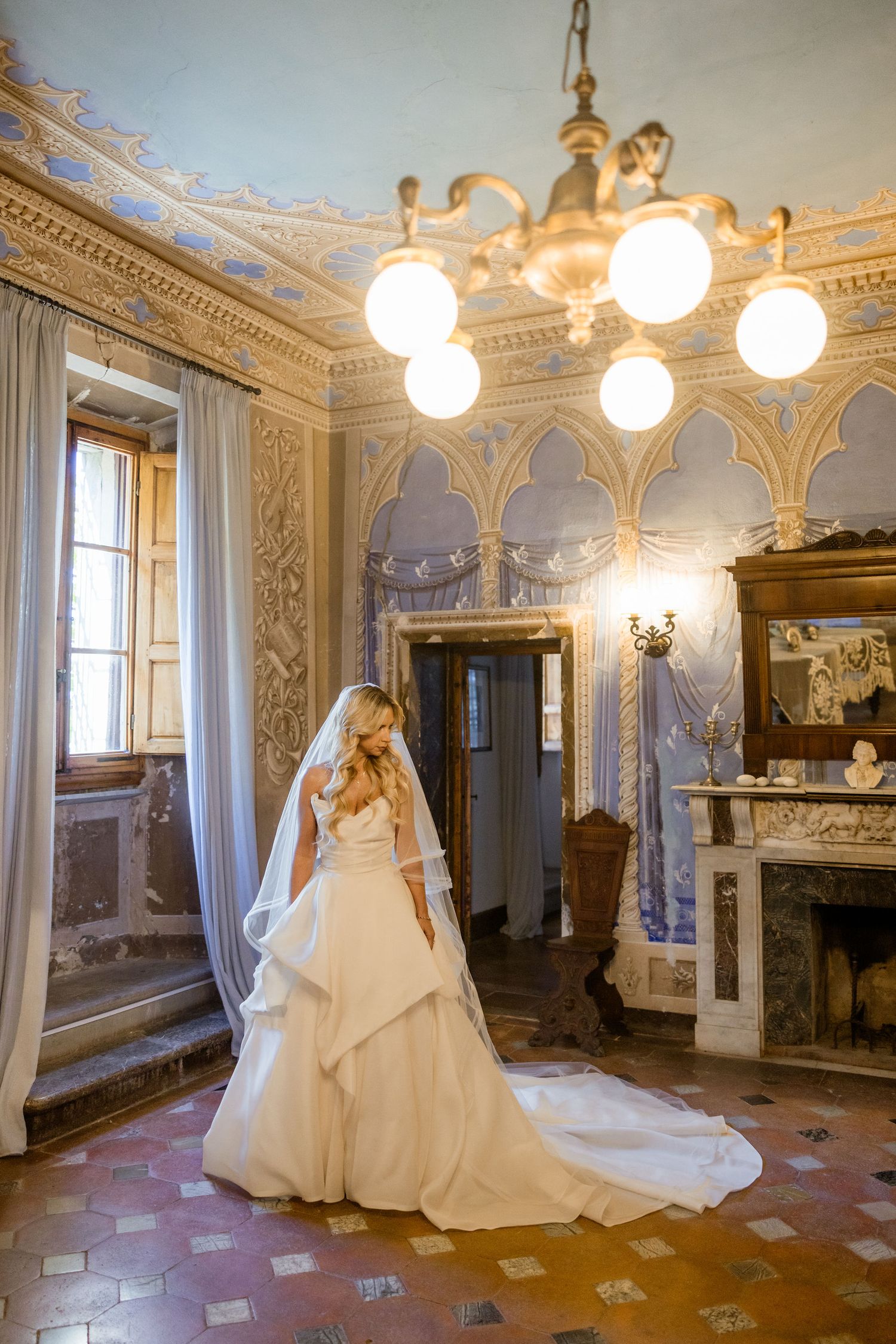A wedding dress displayed in a luxurious ornate room with elegant white walls, decorative moldings and crystal chandeliers.
