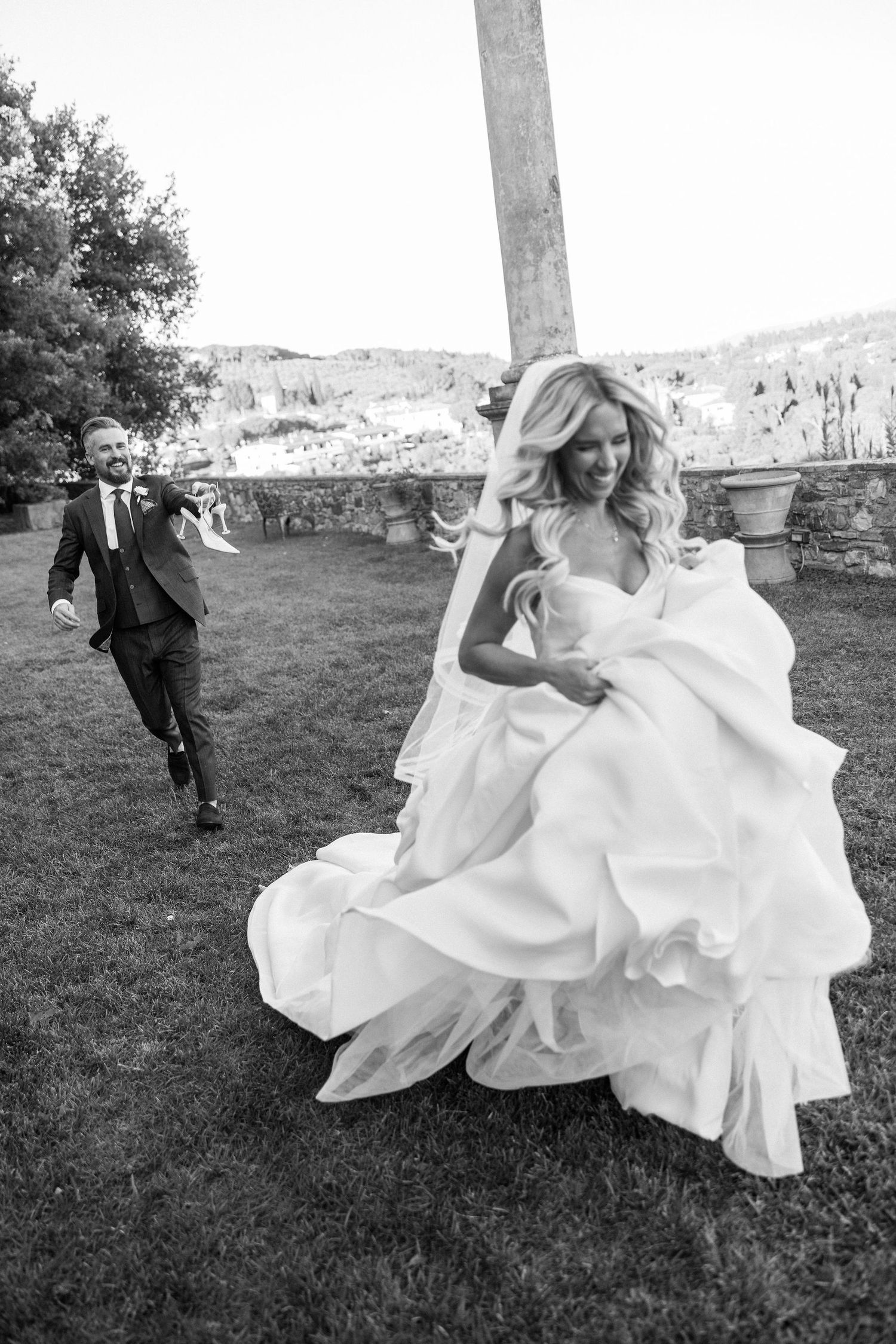 A playful wedding scene shows a bride in a flowing white dress running through a grassy field while someone chases behind.
