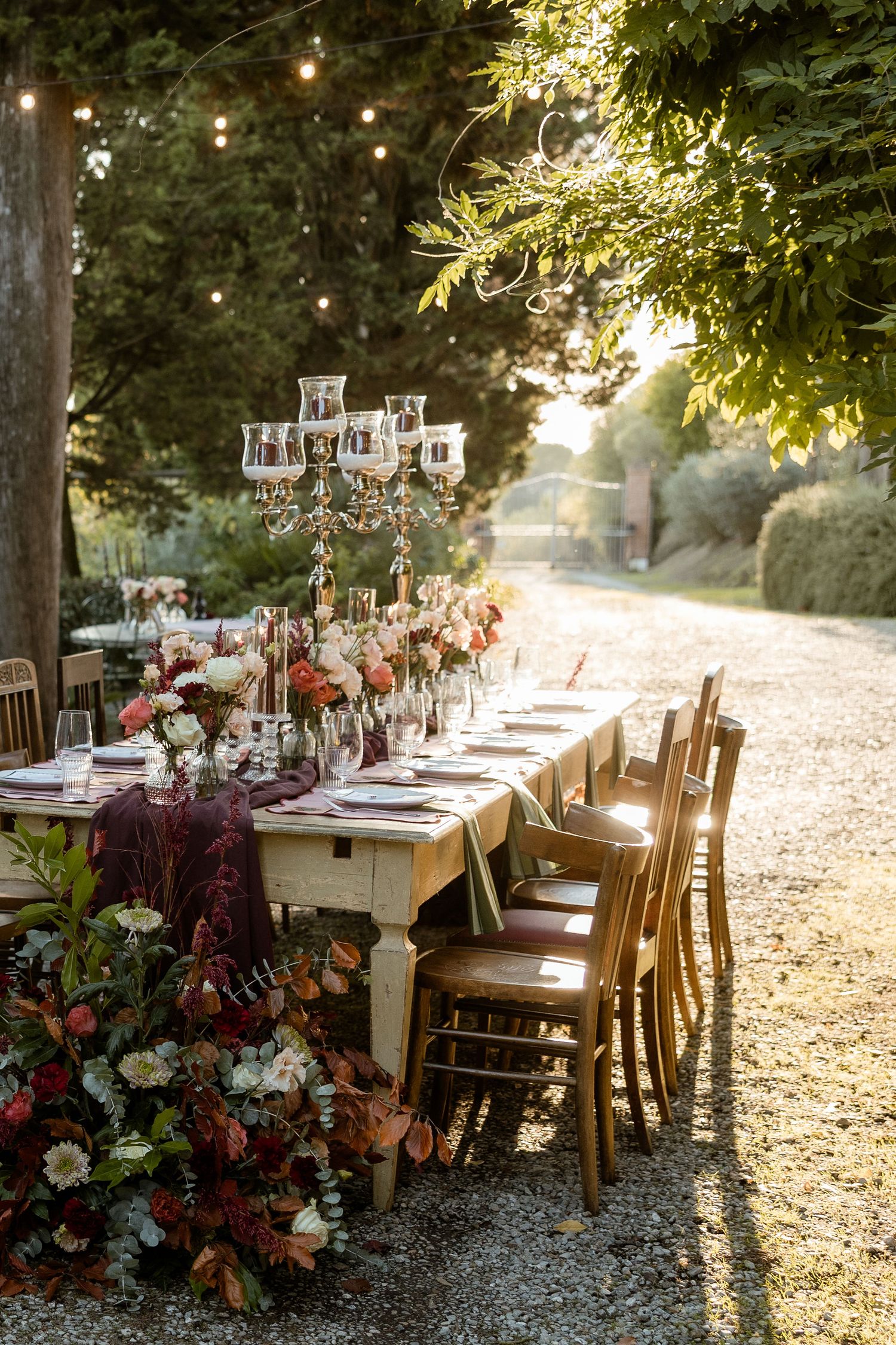 A rustic outdoor wedding table setup with crystal chandeliers, autumn flowers and string lights in a garden path setting.