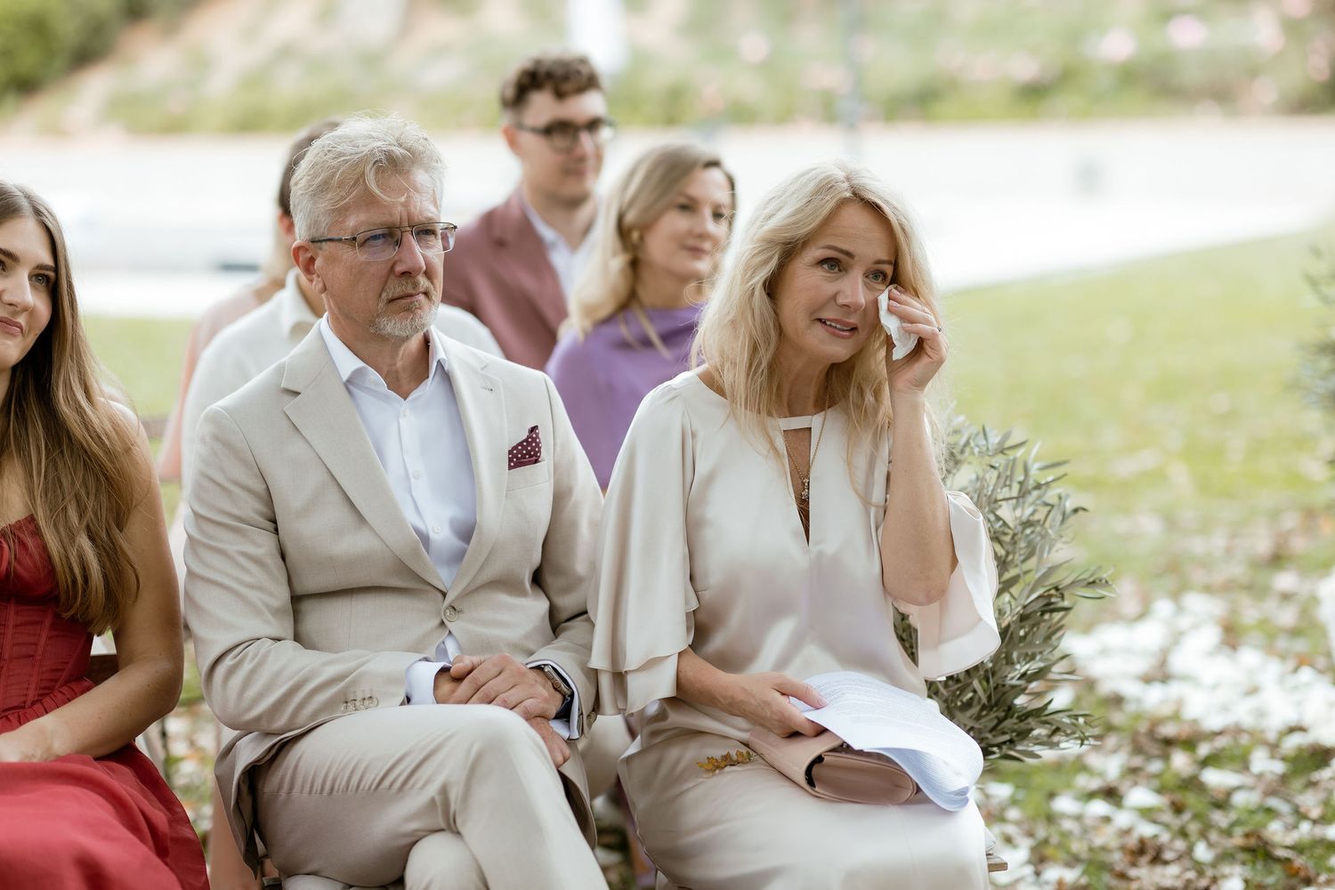 A group of people sit together outdoors in a garden setting with white flowers blooming in the background.