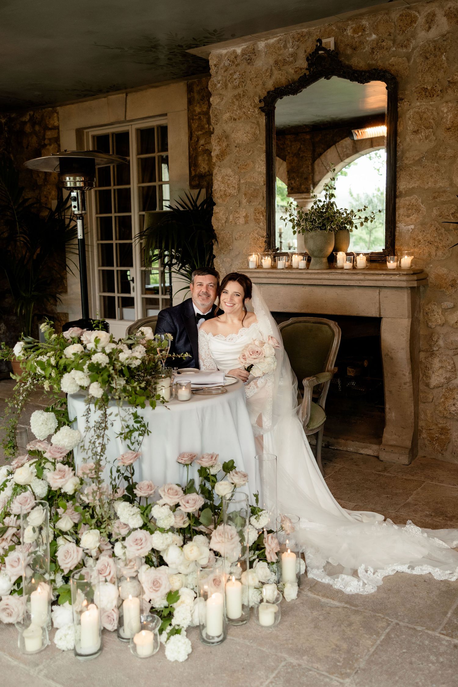 An elegant wedding table arrangement with white flowers and candles set against a rustic stone wall backdrop.