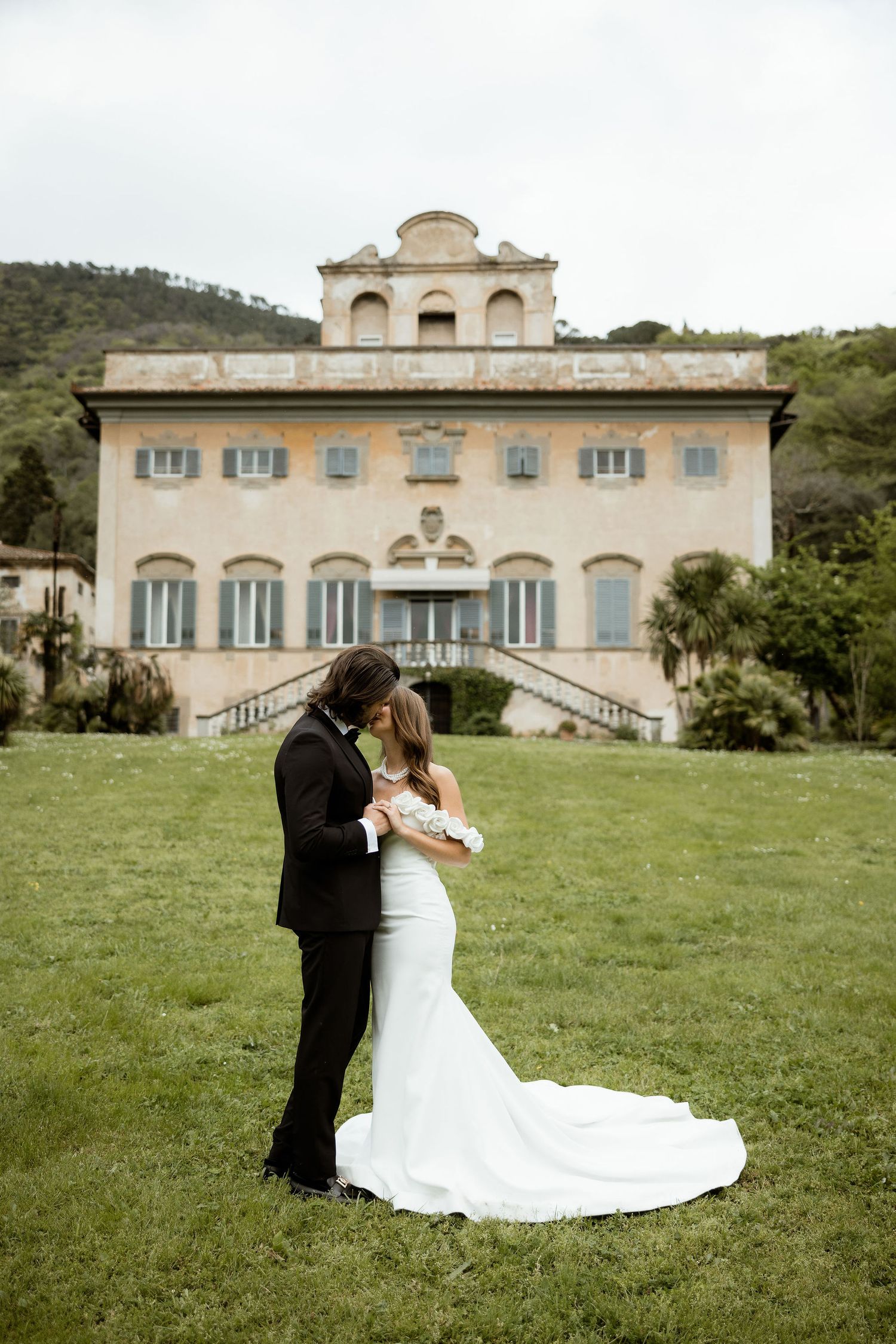 A series of romantic wedding photos set against a beautiful Italian villa with cream-colored walls and blue shutters framed by green lawns.
