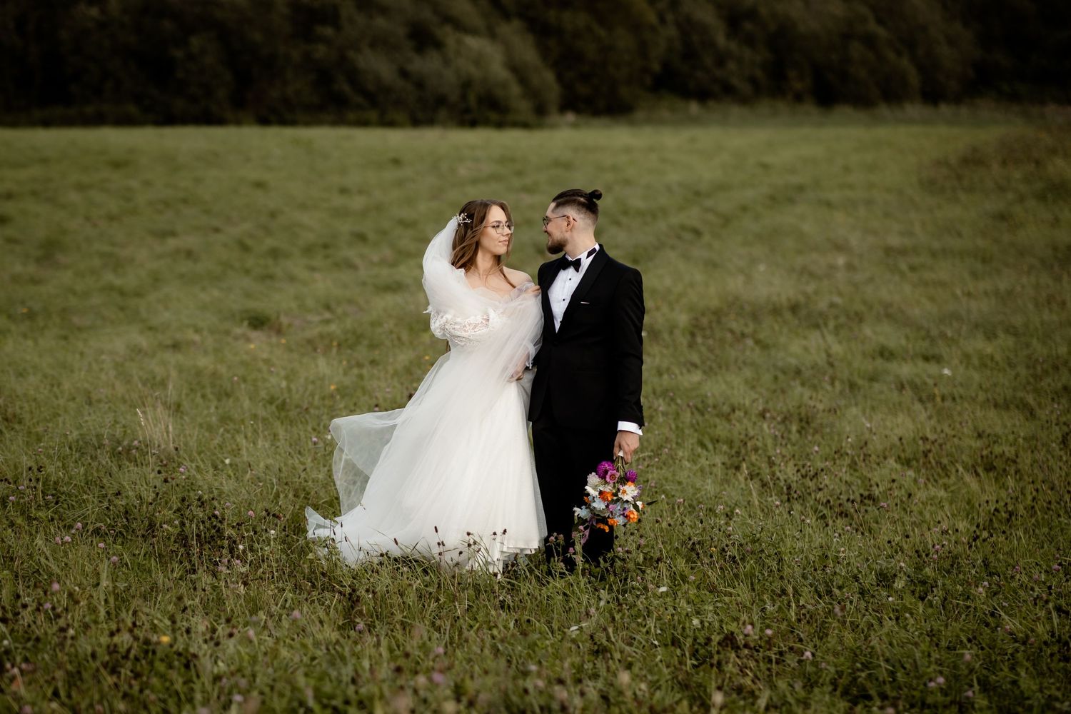 A bride and groom pose together in a scenic green meadow on their wedding day.