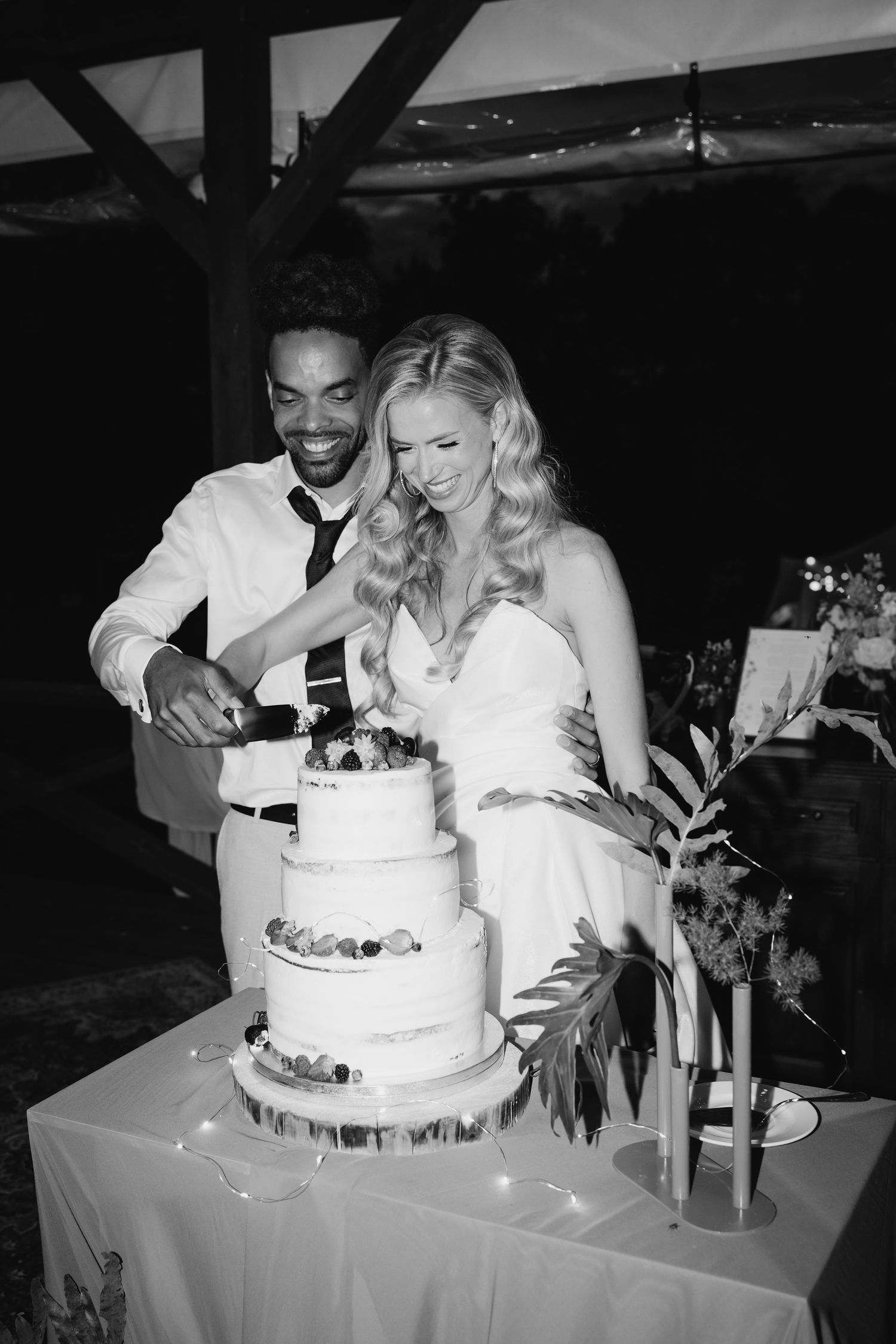 A couple cuts their elegant white tiered wedding cake together at their wedding reception in black and white photography.