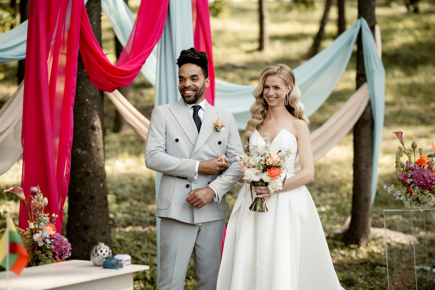 Newlyweds smile together during their forest wedding ceremony decorated with red and blue drapes.