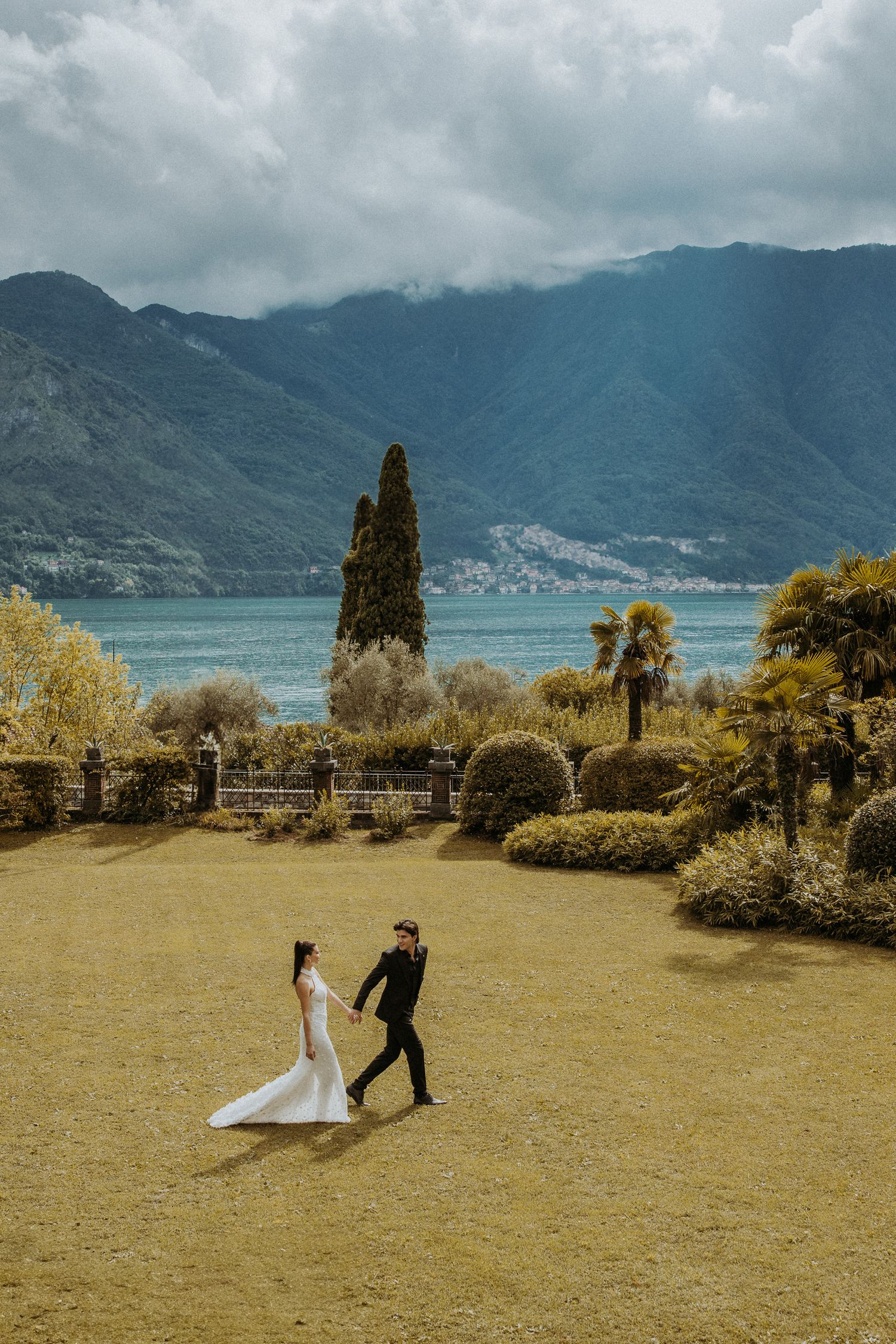 Wedding couple dancing in garden with cypress trees and mountain lake vista.