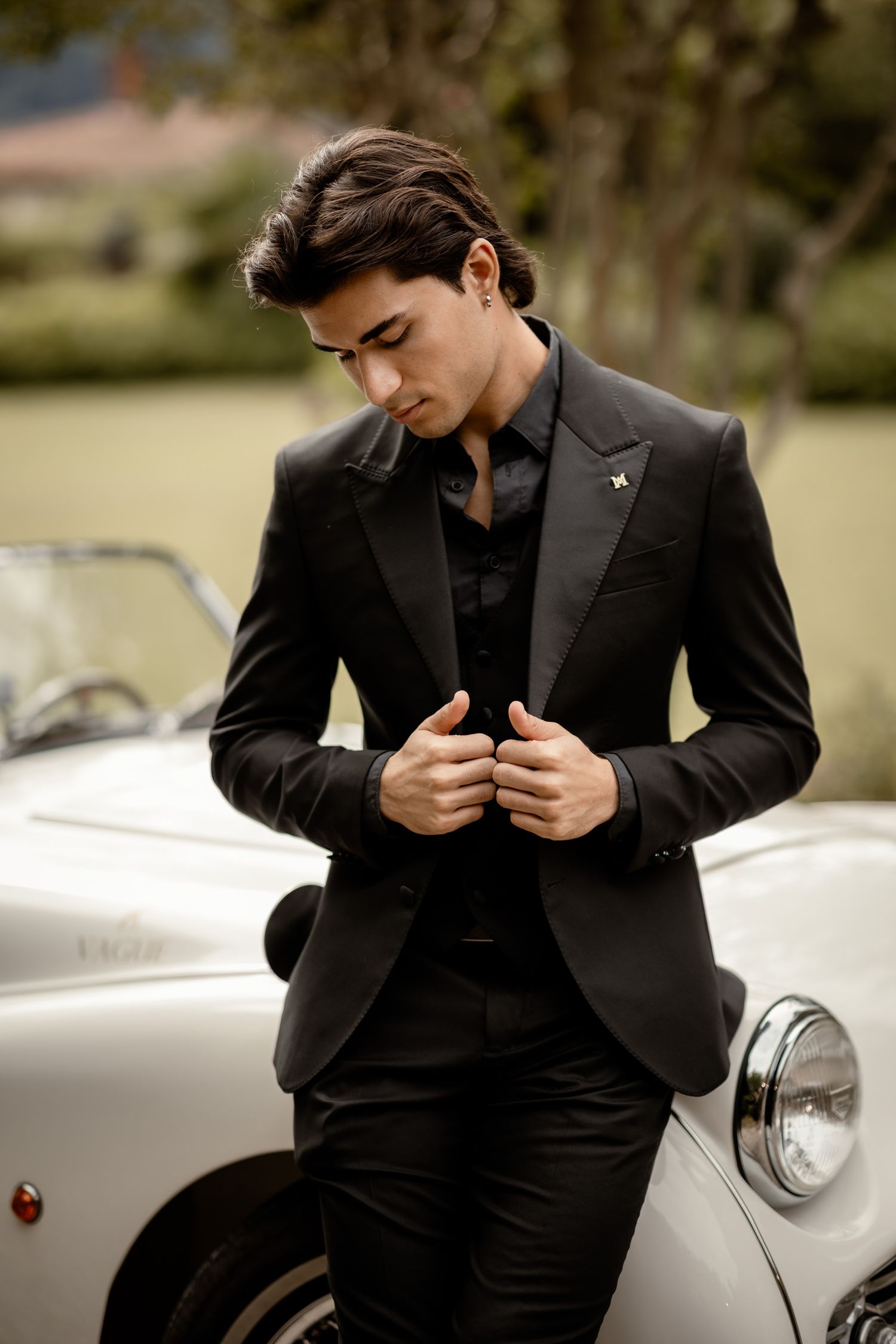 Classic black suit styled against vintage white car in elegant outdoor setting.