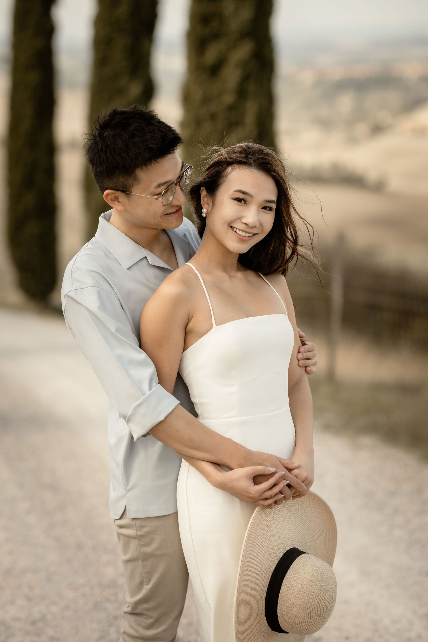 Elegant couple in light summer outfits posing together on cypress-lined path in Tuscany.