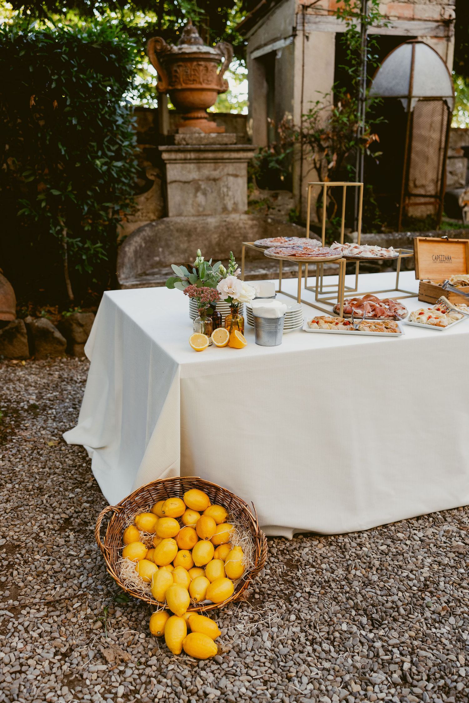 A rustic outdoor buffet table with white linens and scattered citrus fruits on gravel ground near stone pillars.