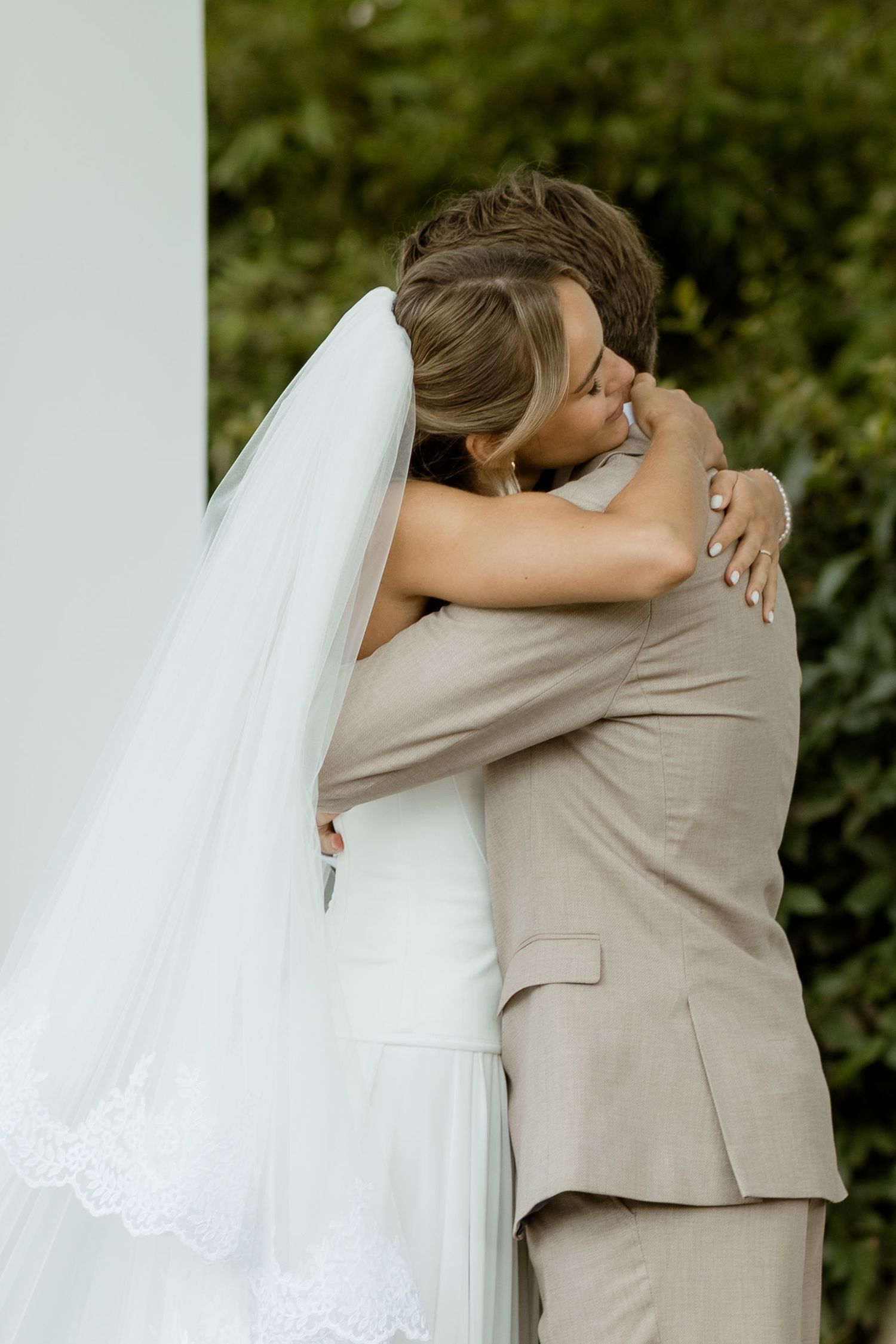 A bride in a white wedding dress and groom in beige suit embrace lovingly in an outdoor garden setting.