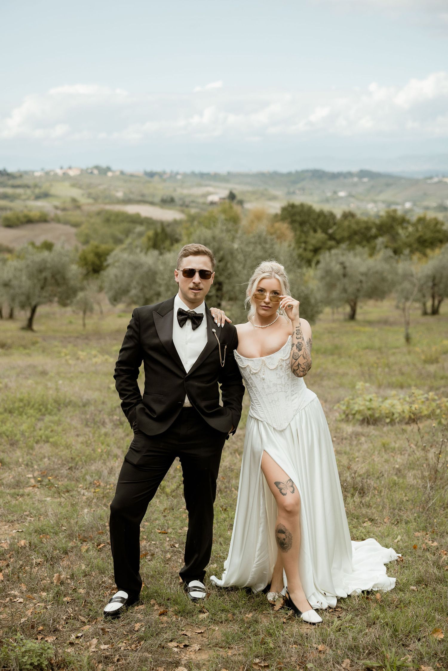 A couple poses in wedding attire on a scenic hilltop with olive trees and rolling countryside in the background.