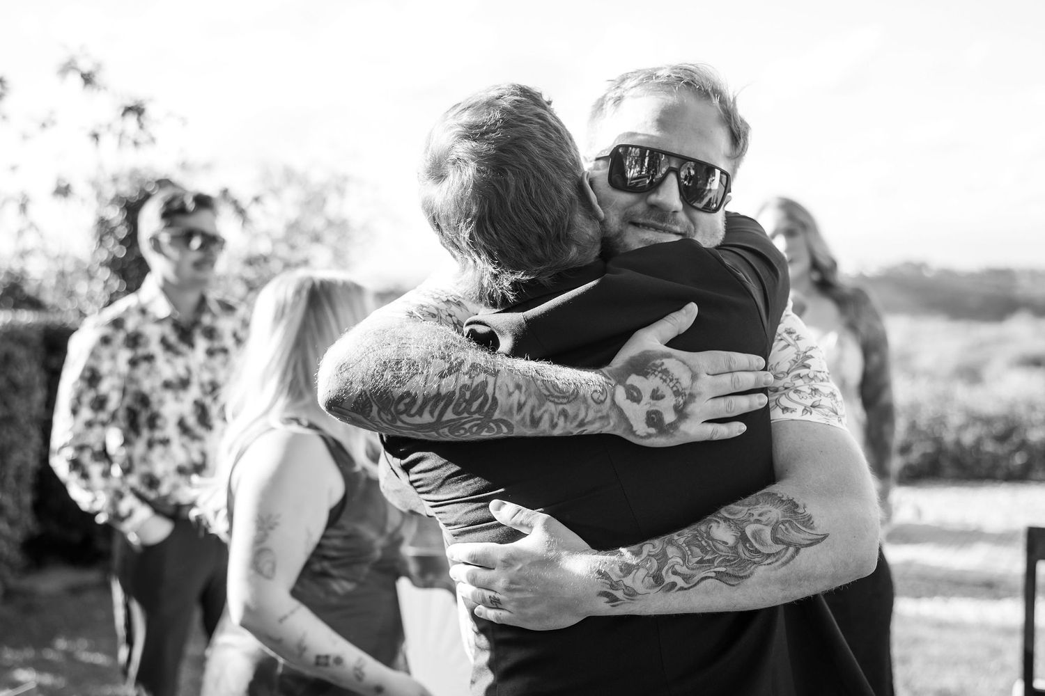 Two people sharing an emotional embrace outdoors in black and white with visible tattoos.