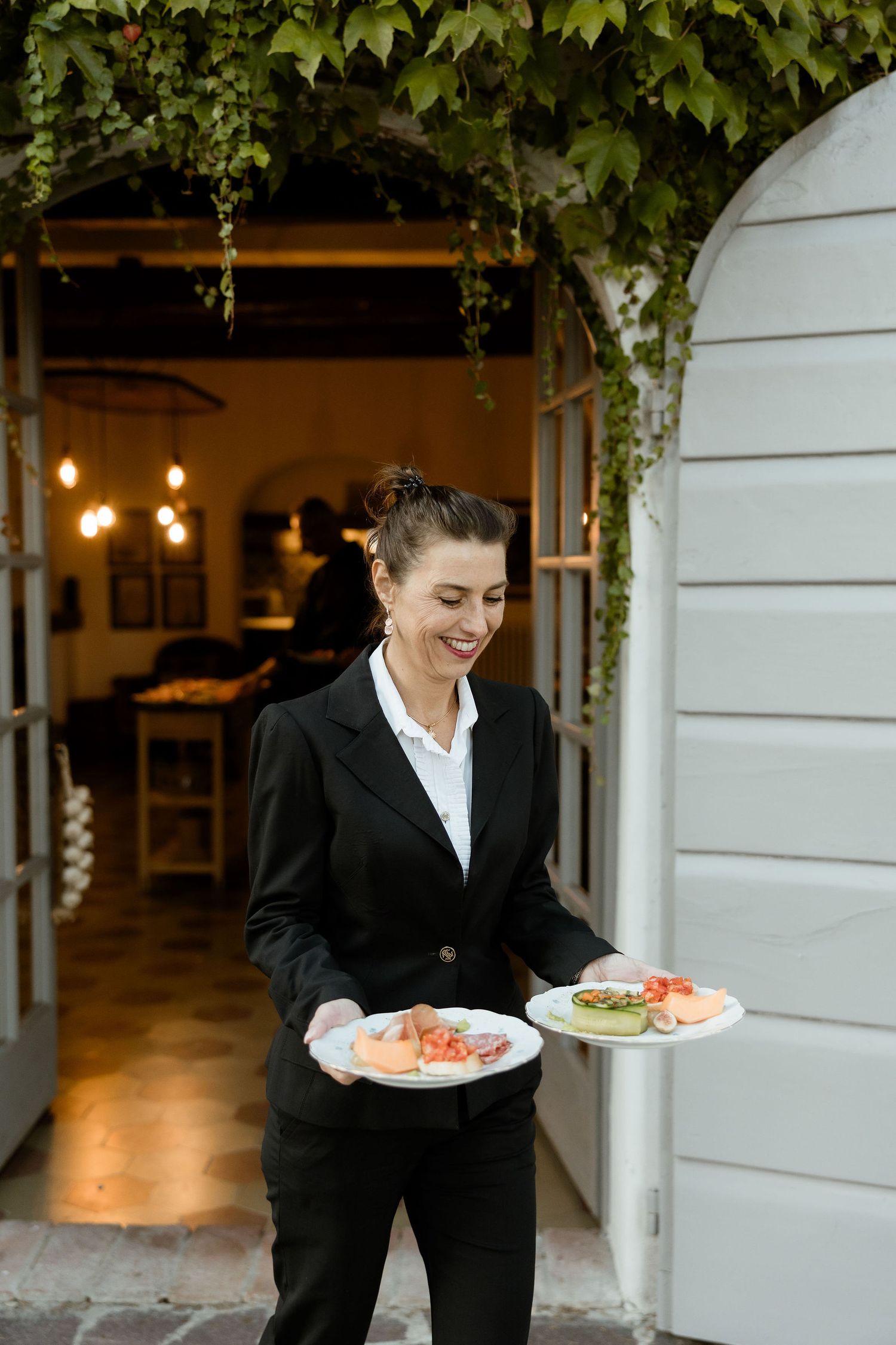 Wait staff in black uniforms serve plates of food at an outdoor venue with white walls and hanging greenery.