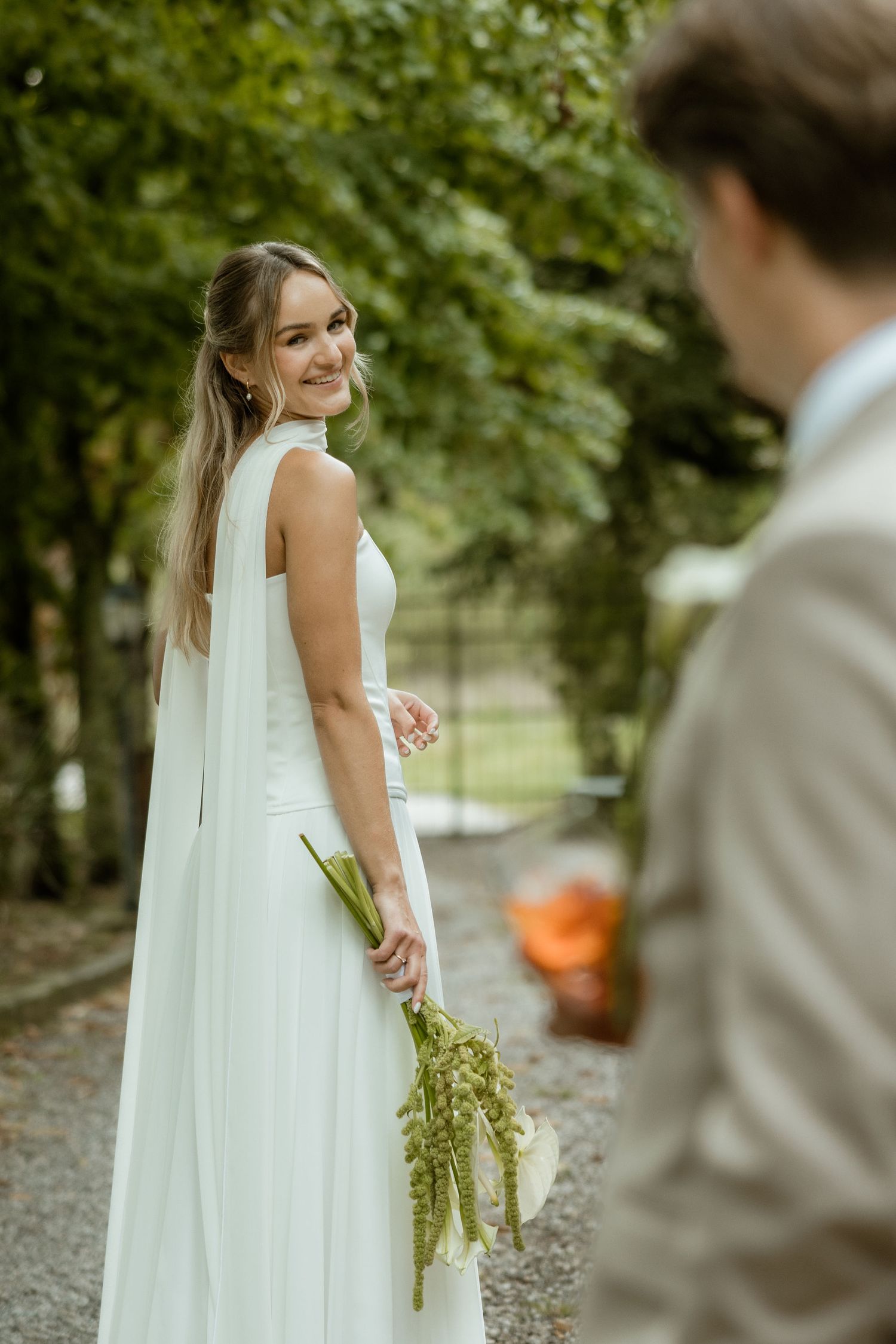 A bride in a flowing white wedding dress poses outdoors while being photographed in a garden setting.