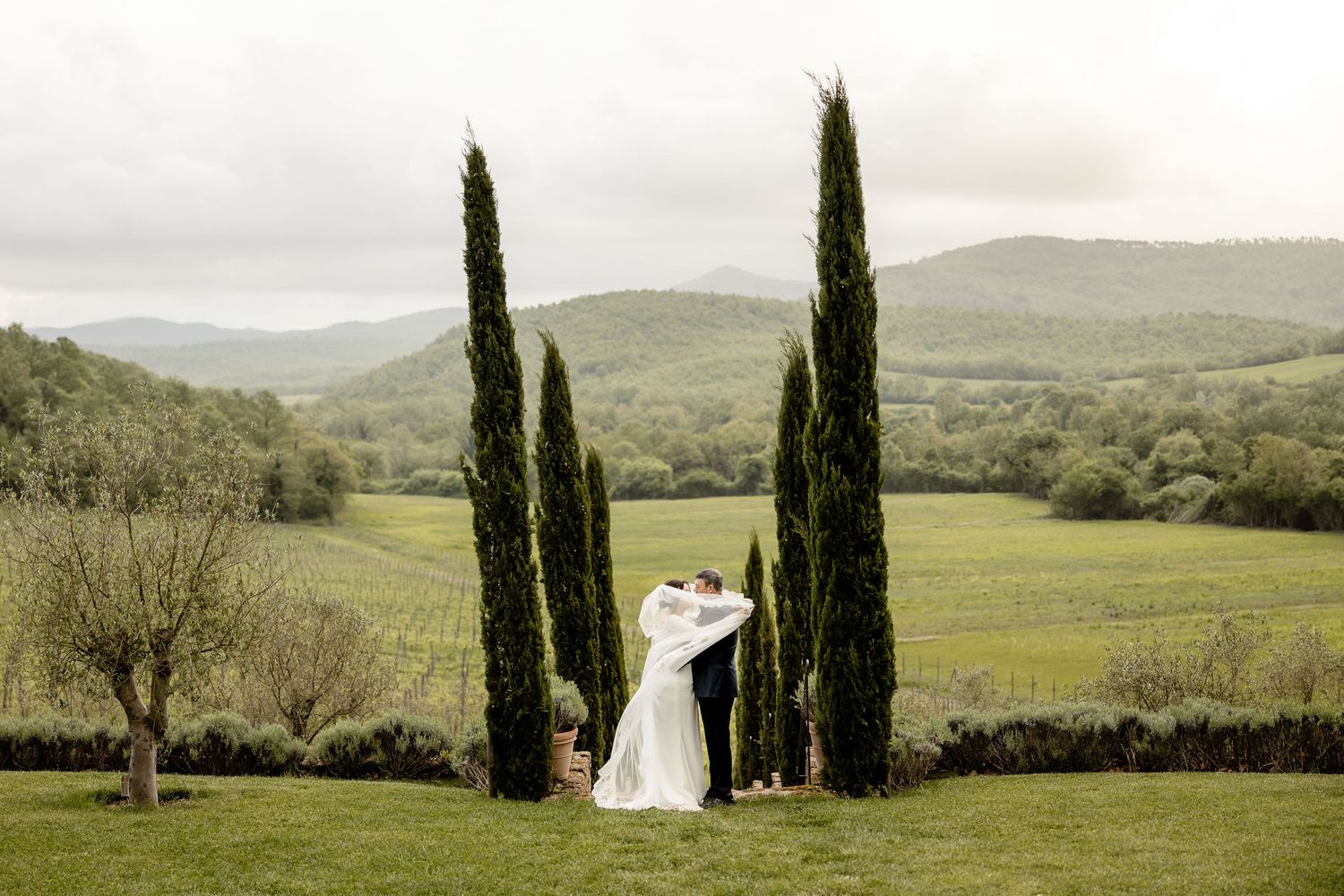 A romantic wedding pose between towering cypress trees overlooking rolling hills in the Tuscan countryside.
