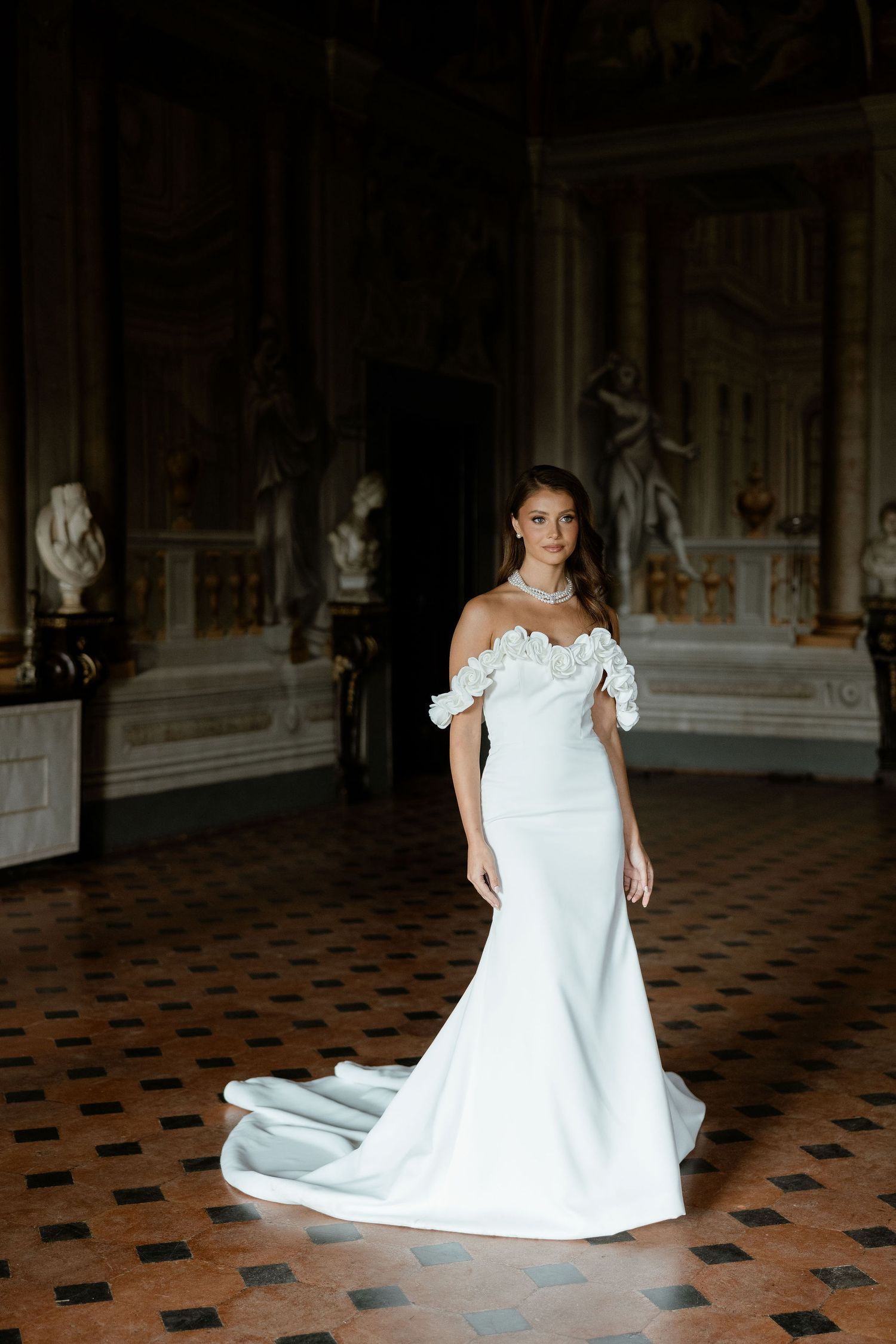 Elegant off-shoulder white wedding gown with fitted silhouette and flowing train photographed in ornate historic interior.