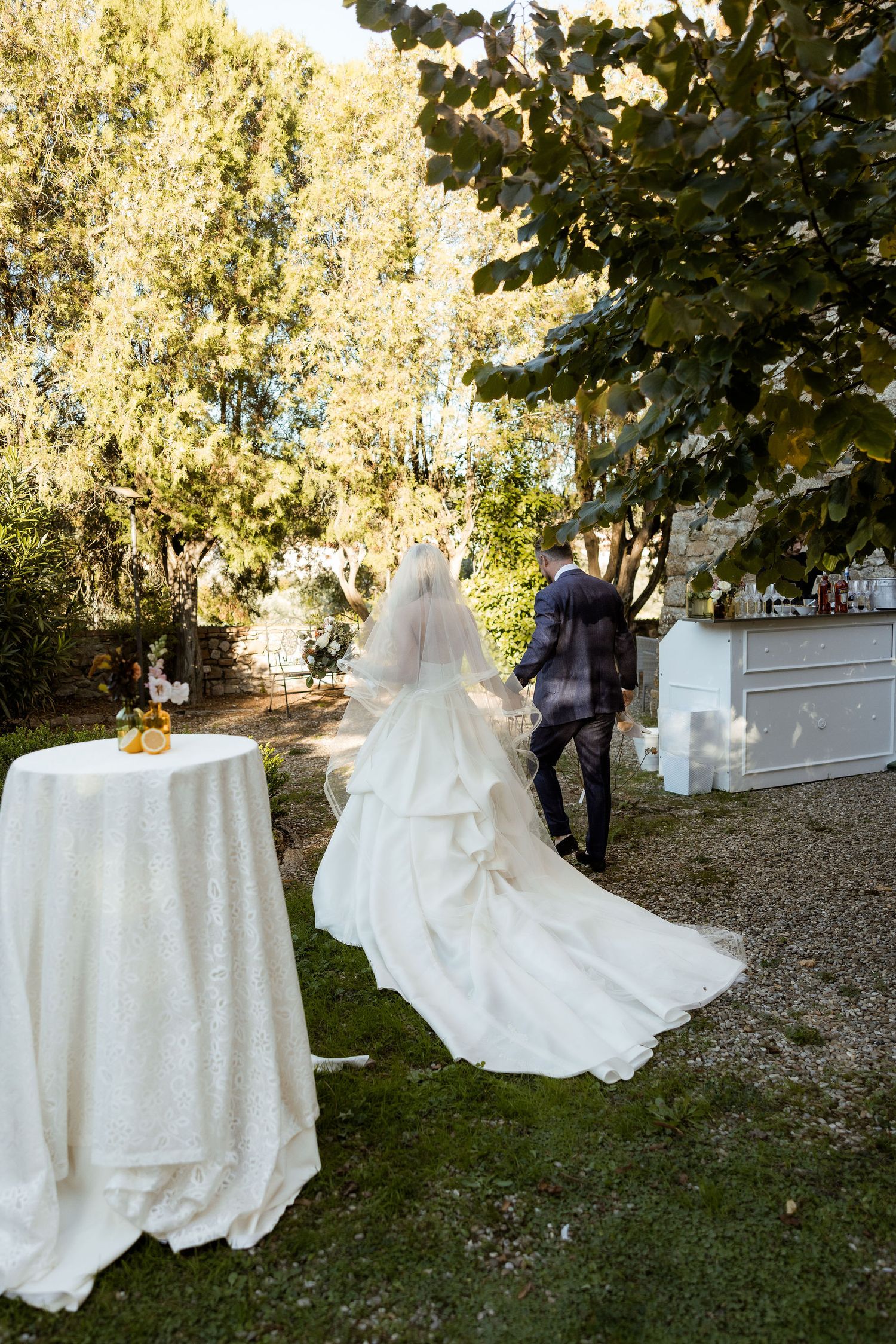 A series of white wedding dresses and tables arranged outdoors in a garden setting with trees and gravel paths.