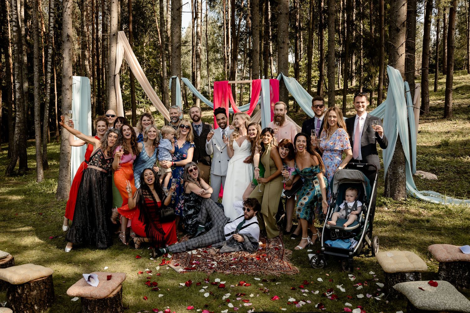 Large group wedding photo with colorful decorations and fabric backdrops in forest setting.