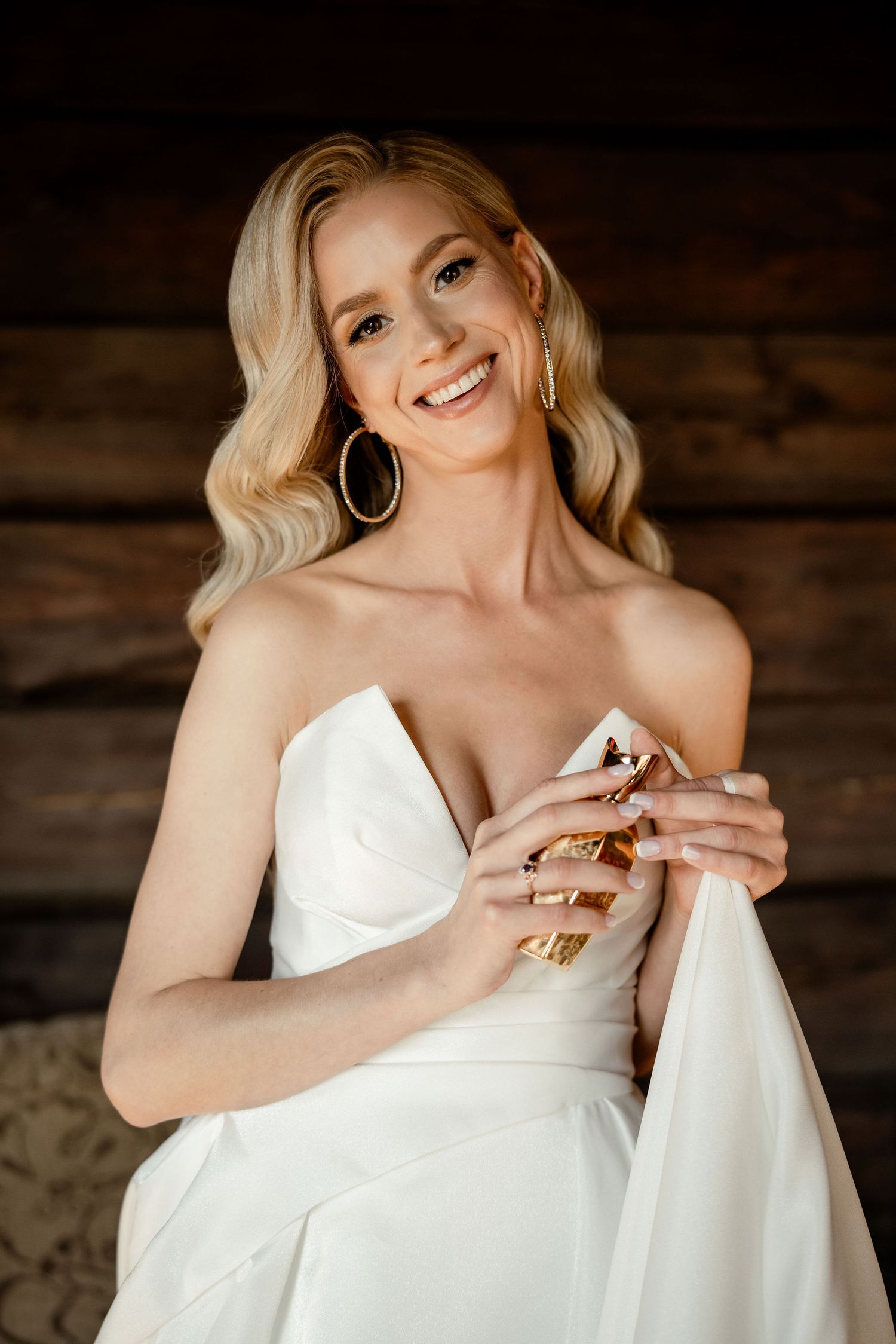 A portrait in a wedding dress with blonde wavy hair and hoop earrings against a rustic wooden backdrop.