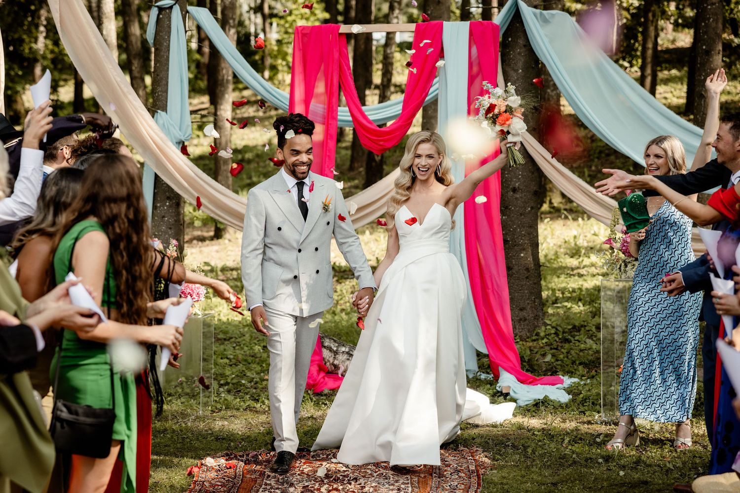 Wedding celebration in forest with guests throwing petals as couple walks under draped fabric decor.