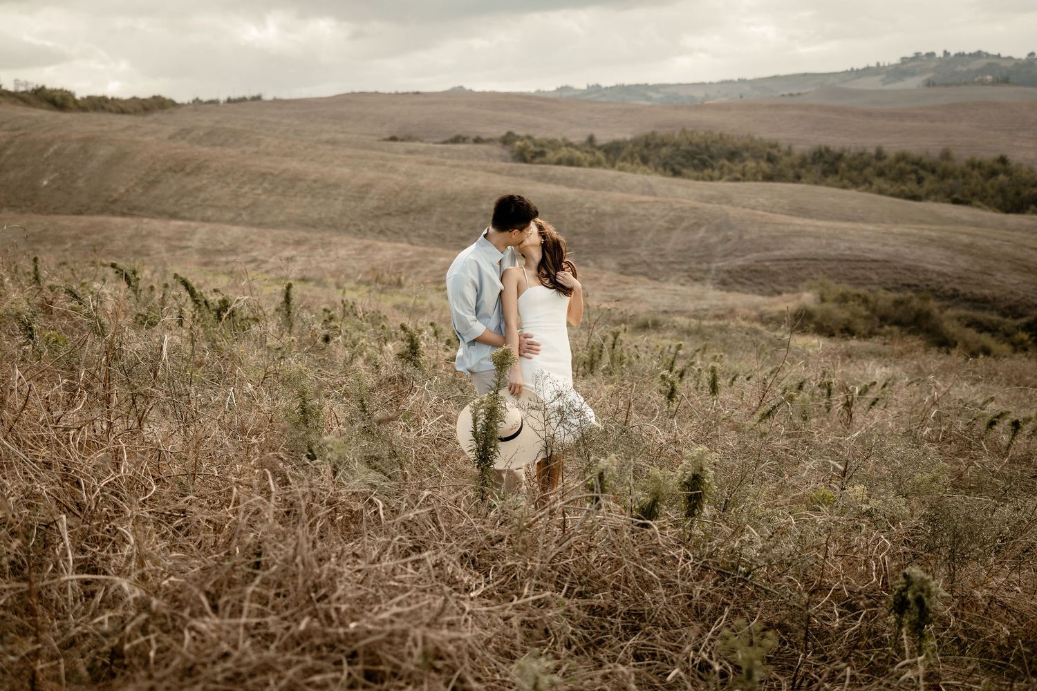 A couple in white stands among dry grass with rolling hills in the background.