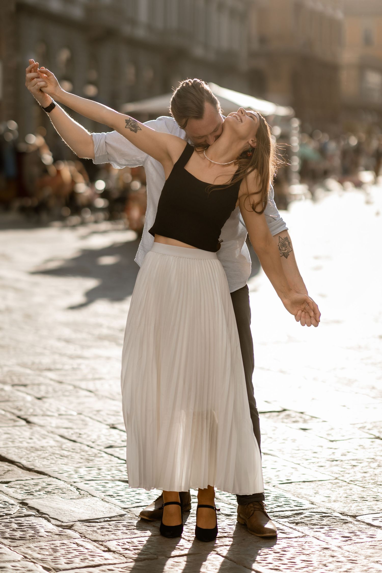 A couple dances intimately on a sunlit cobblestone street, with the woman wearing a flowing white skirt and black top.