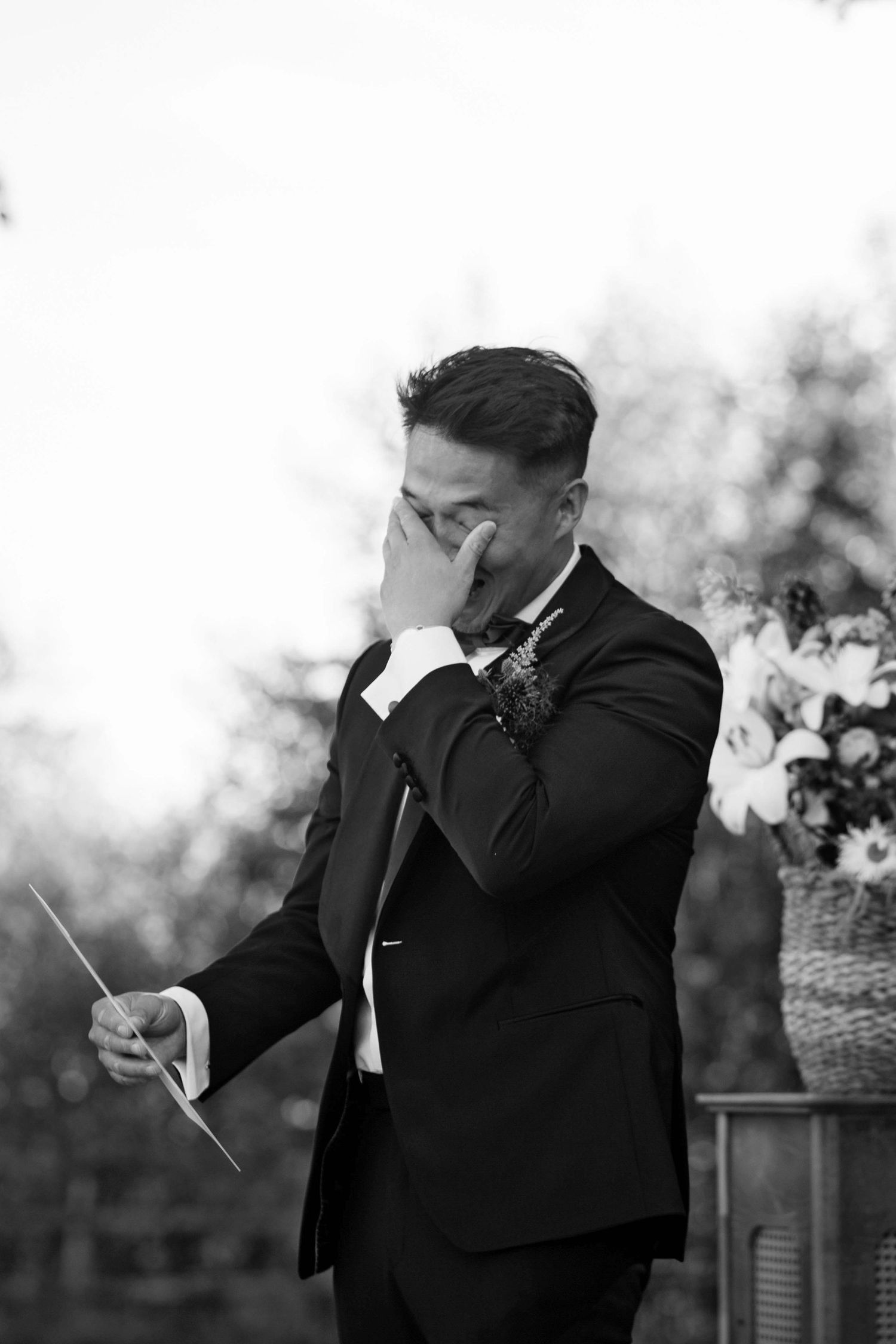 Black and white emotional moment captured as groom in dark suit wipes tears during outdoor wedding ceremony.