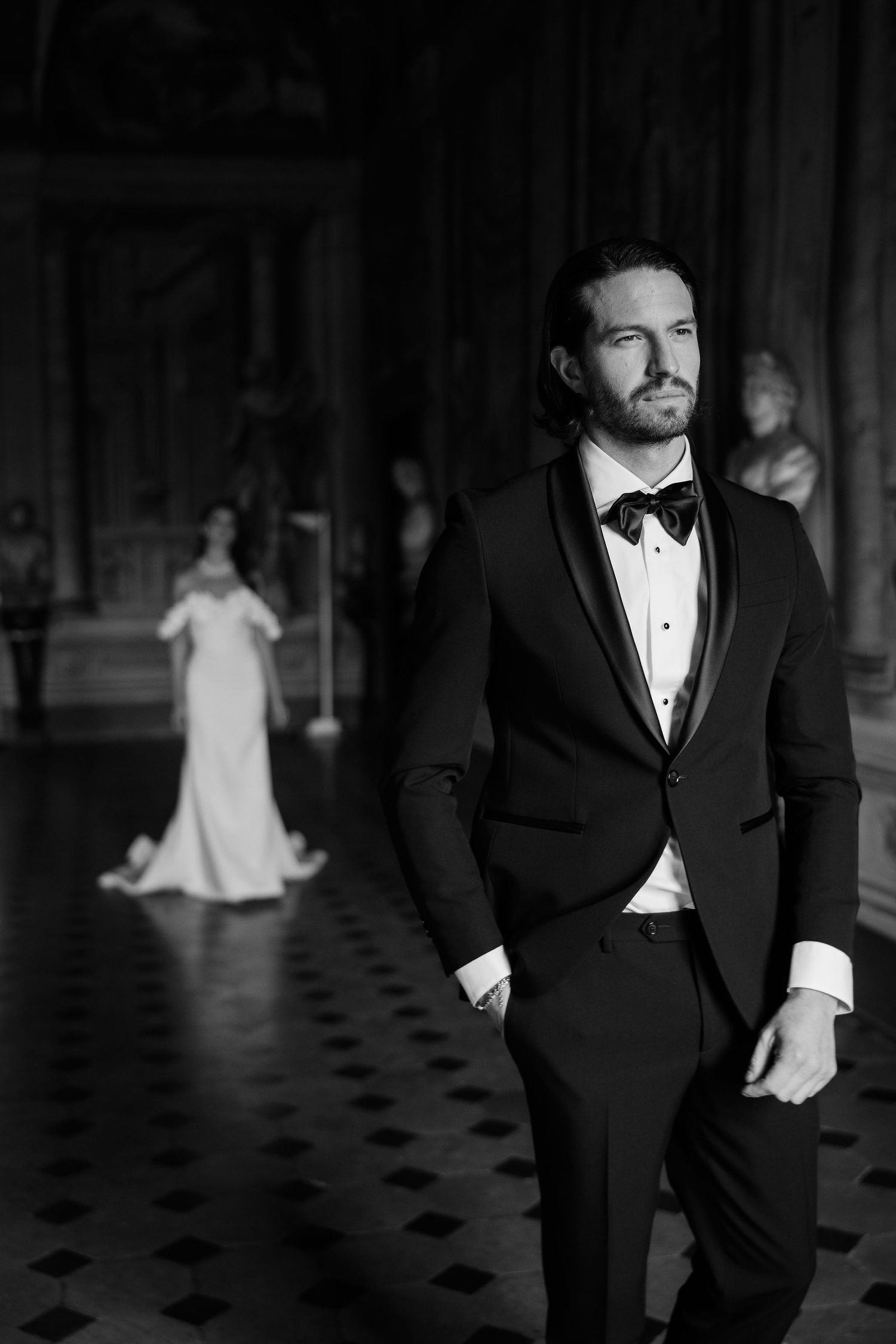 A groom in a black tuxedo and bowtie poses in an elegant venue while a bride in white stands in the background.