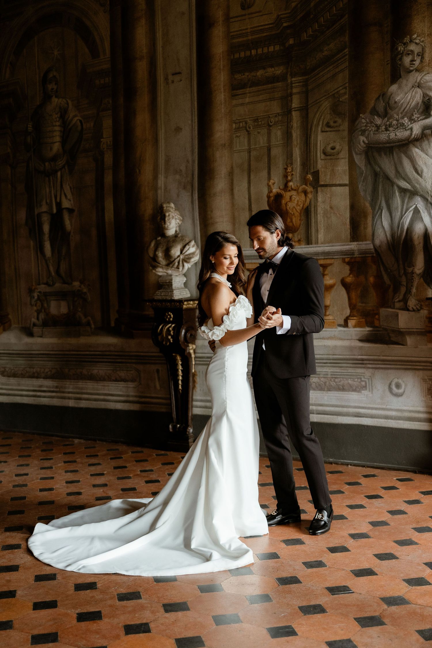 A couple poses in an elegant historic setting with ornate marble sculptures and tiled floors during their wedding day.