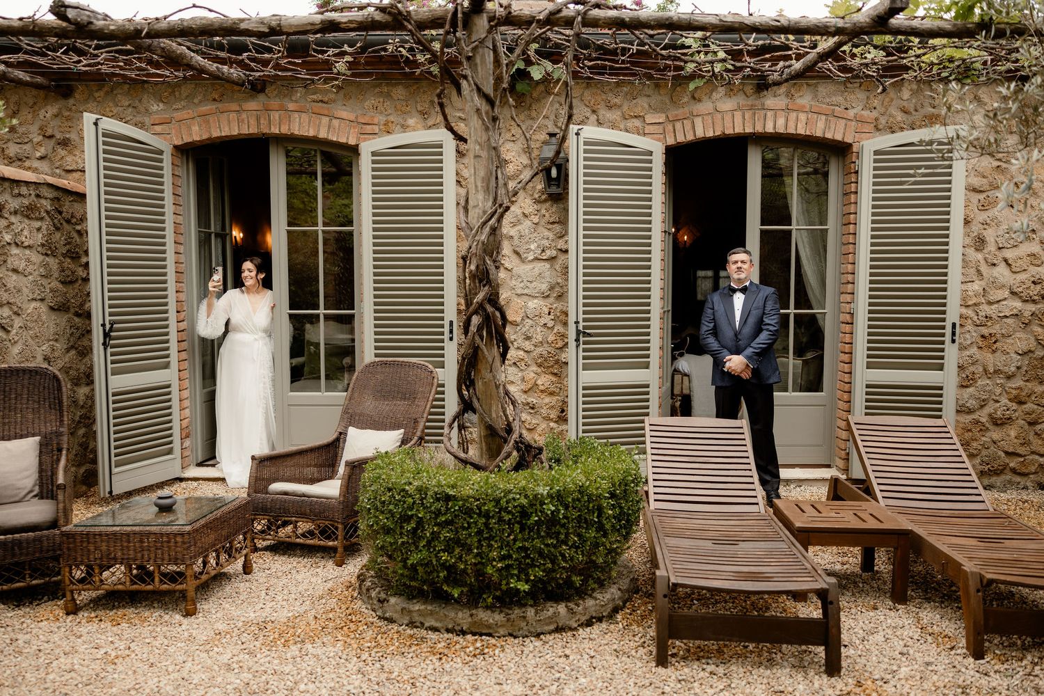 Two figures standing in separate doorways of a rustic stone building with lounge chairs and manicured shrubs in courtyard.