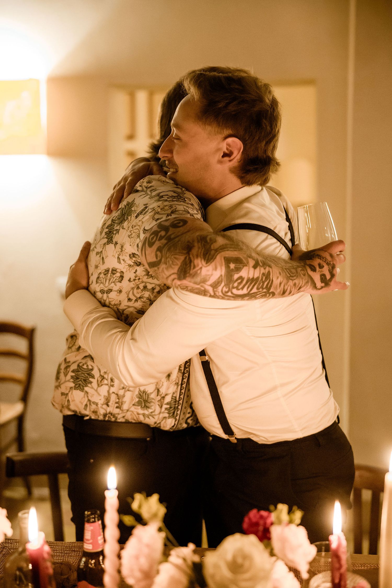 A couple embraces at a candlelit dinner table with soft lighting and pink flowers creating a romantic atmosphere.