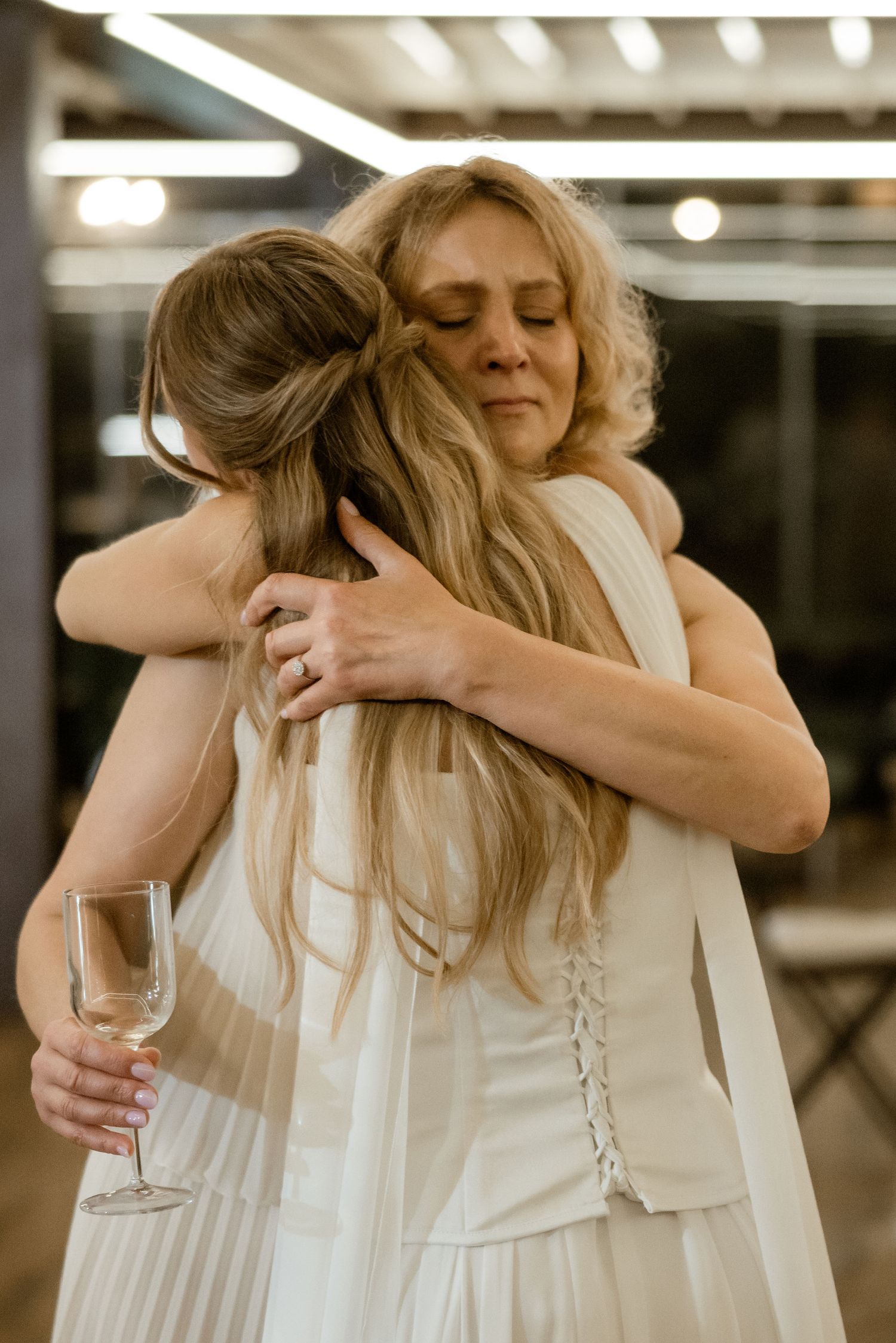 Two people in white dresses share an emotional embrace during what appears to be a special celebration.