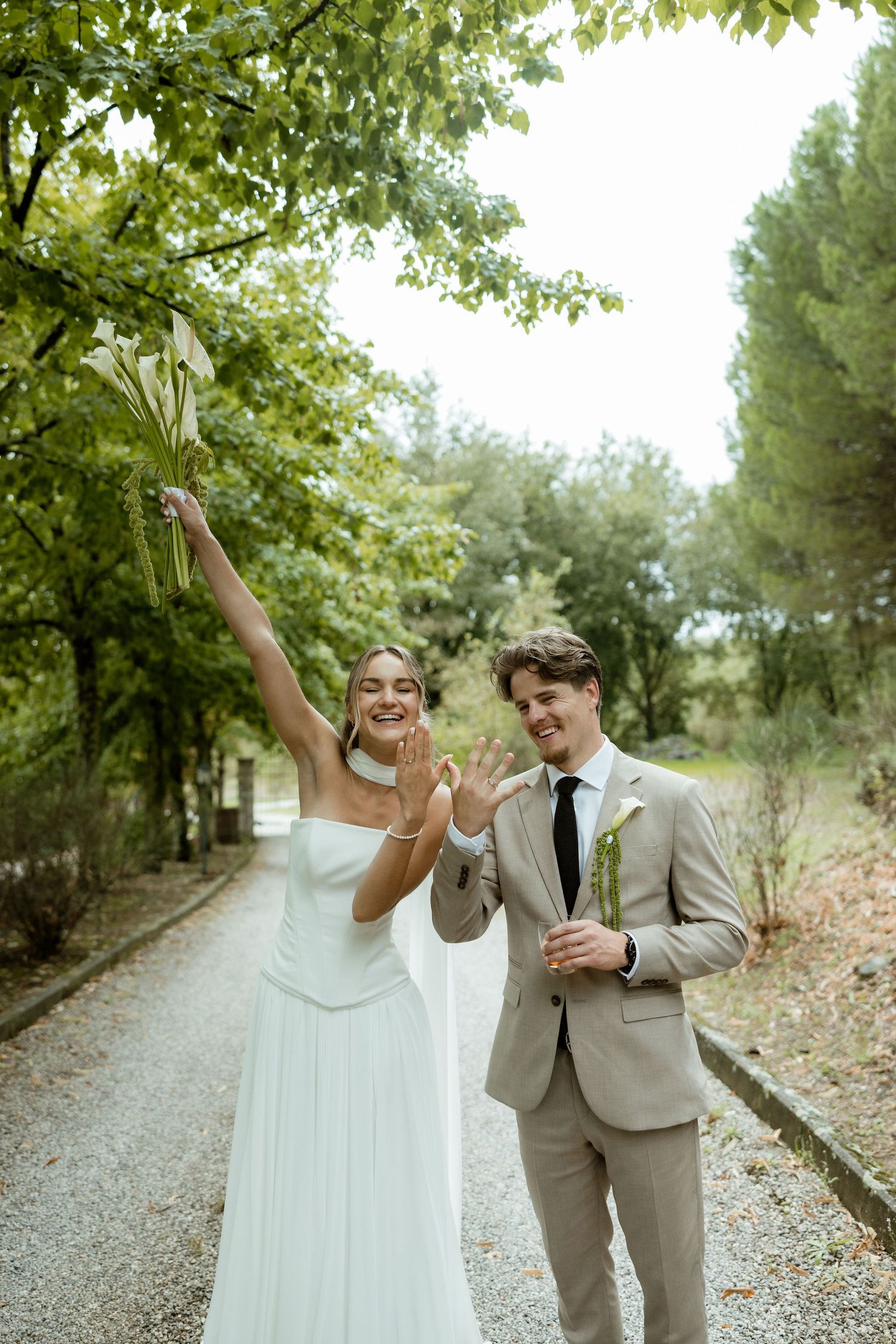 A couple in wedding attire celebrate joyfully on a tree-lined gravel path during their outdoor celebration.