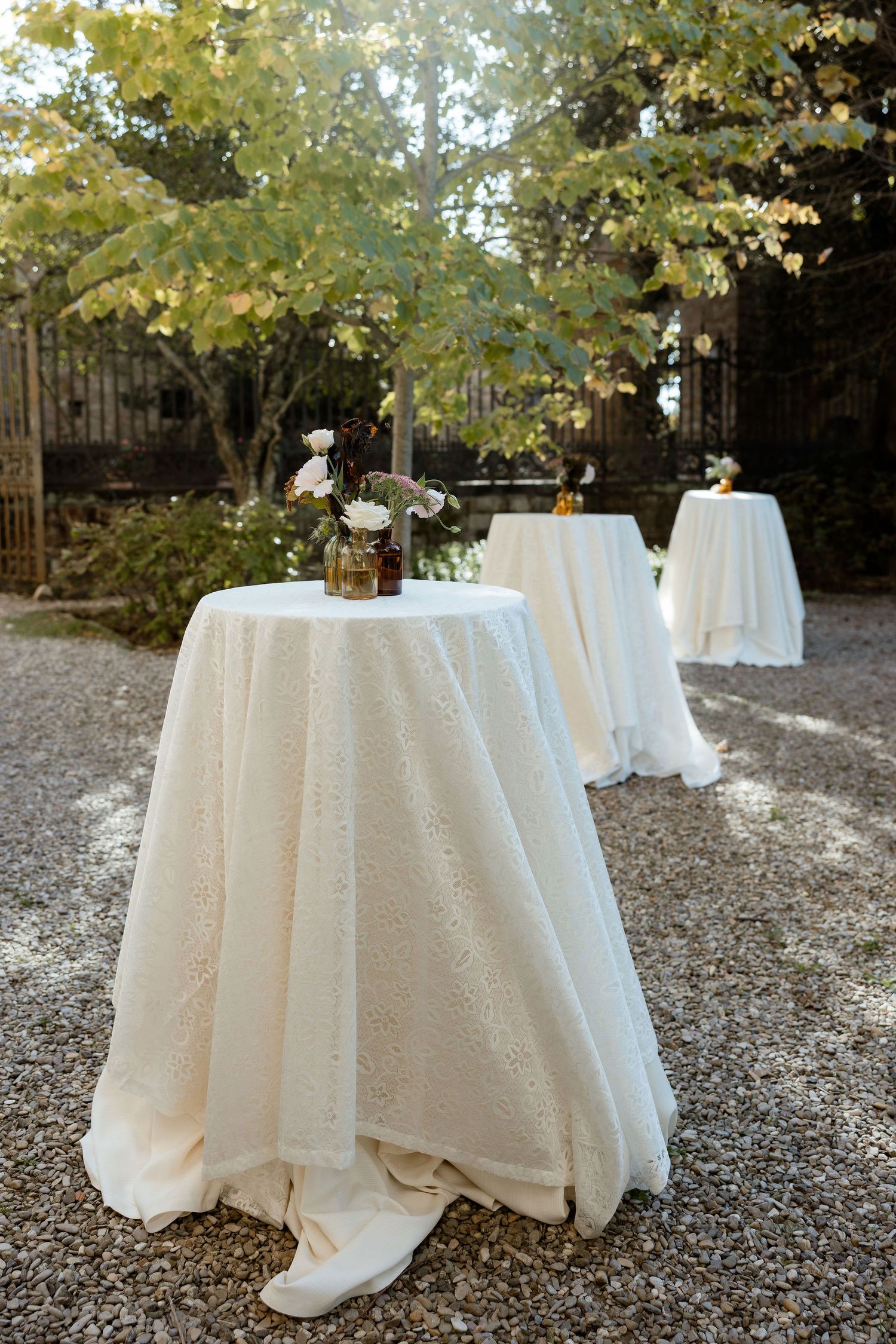 Three round cocktail tables with white floor-length tablecloths set up outdoors under autumn trees for an elegant event.
