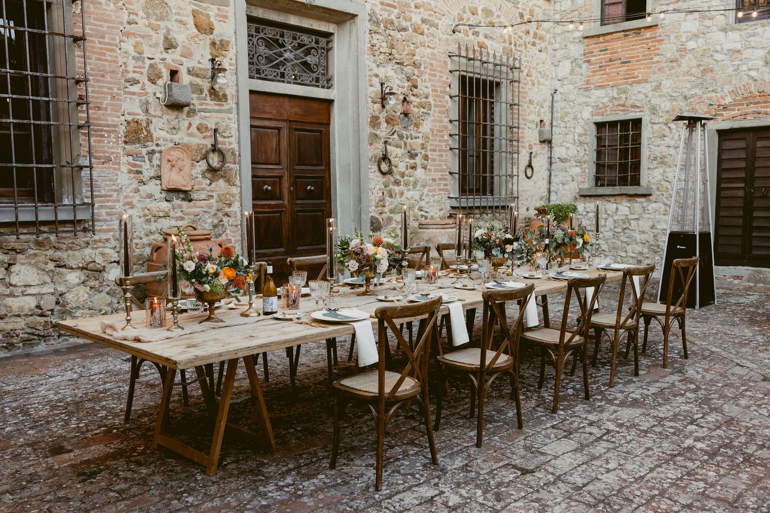 Long wooden tables with white linens and floral centerpieces set up for outdoor dining against rustic stone walls.